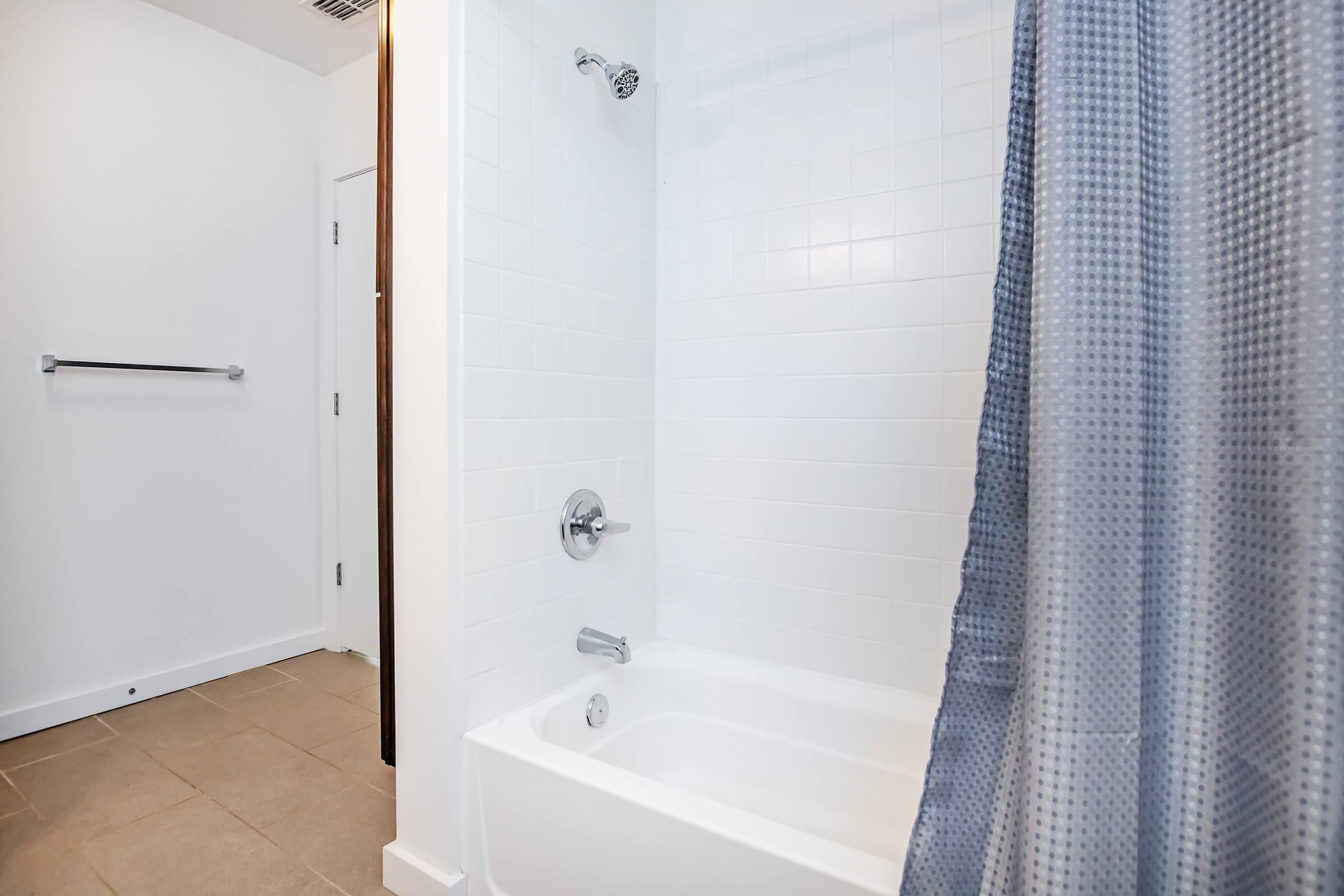 A clean, modern bathroom featuring a tub with a shower head and a gray shower curtain. The walls are white with light-colored tiles on the floor. A towel bar is mounted on the wall, and there is a closed white door leading to another room. The overall ambiance is bright and minimalist.