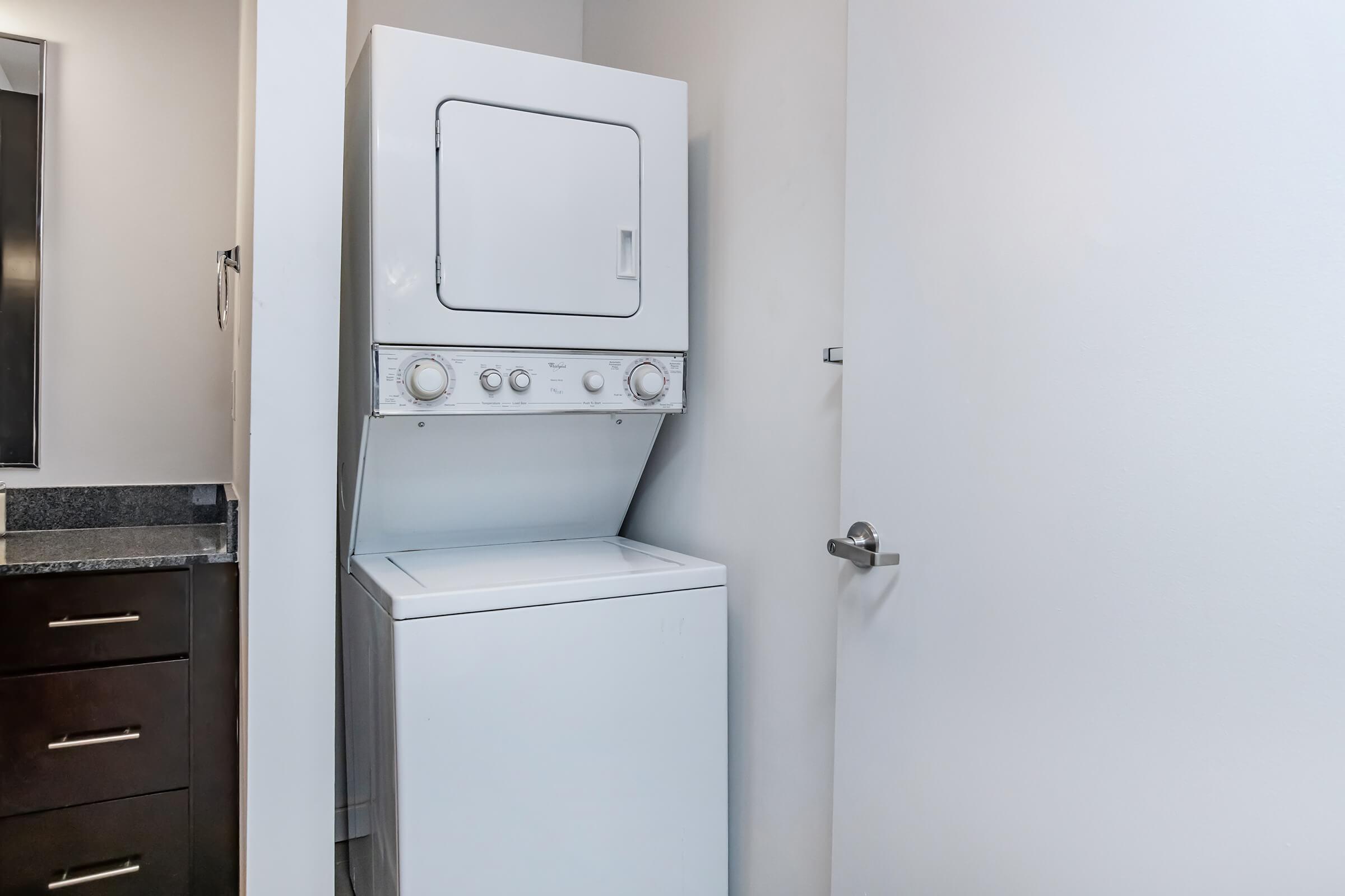 A compact stacked washer and dryer unit in a modern laundry space, nestled against a wall with cabinetry below. The interior features a clean and minimalistic design, with gray walls and a small door nearby. The appliances are predominantly white, adding to the contemporary aesthetic.