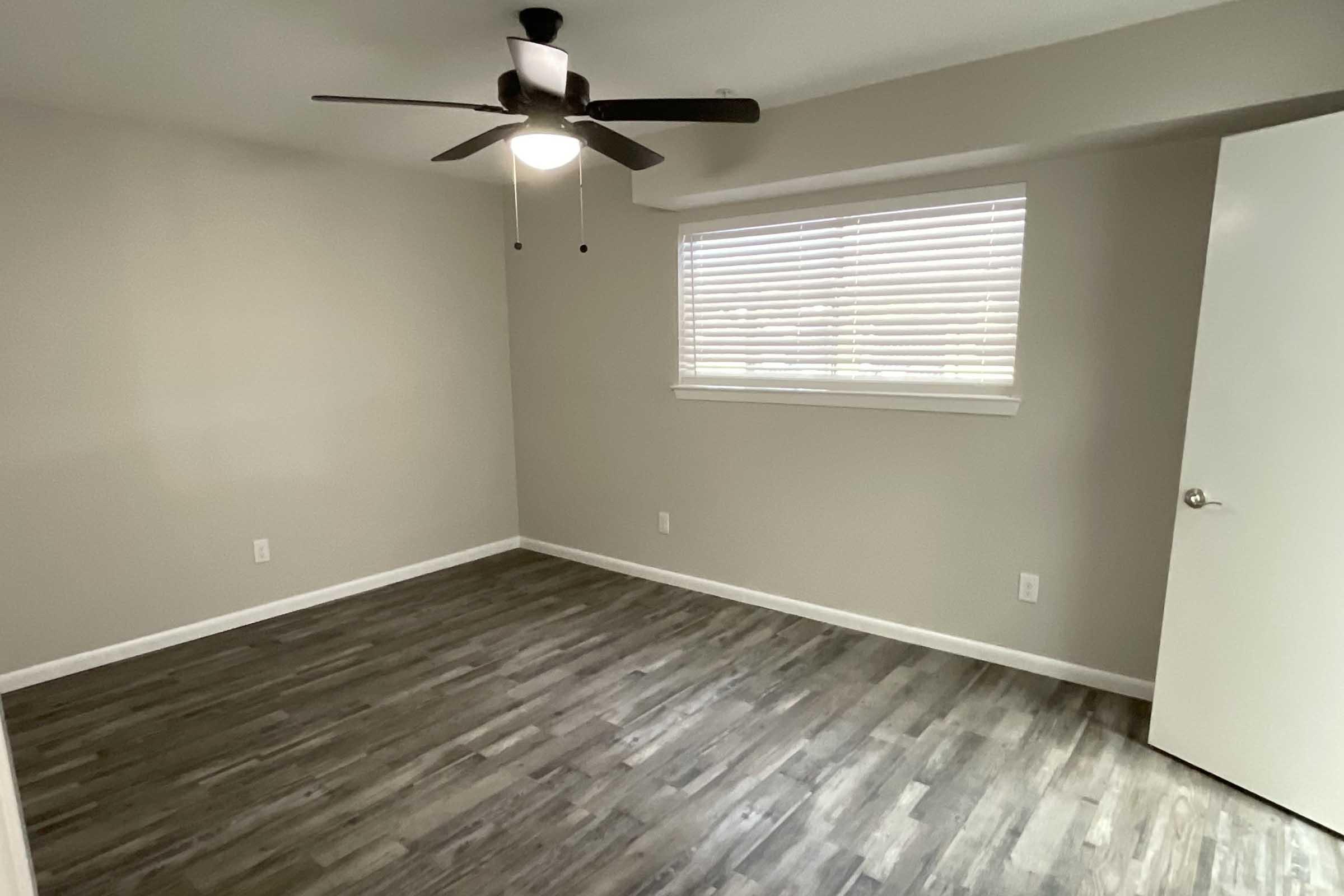 A well-lit empty room featuring a ceiling fan, a window with blinds, and light-colored walls. The floor is covered with modern laminate flooring, and there is a door on the right side of the image. The overall aesthetic is clean and minimalist.