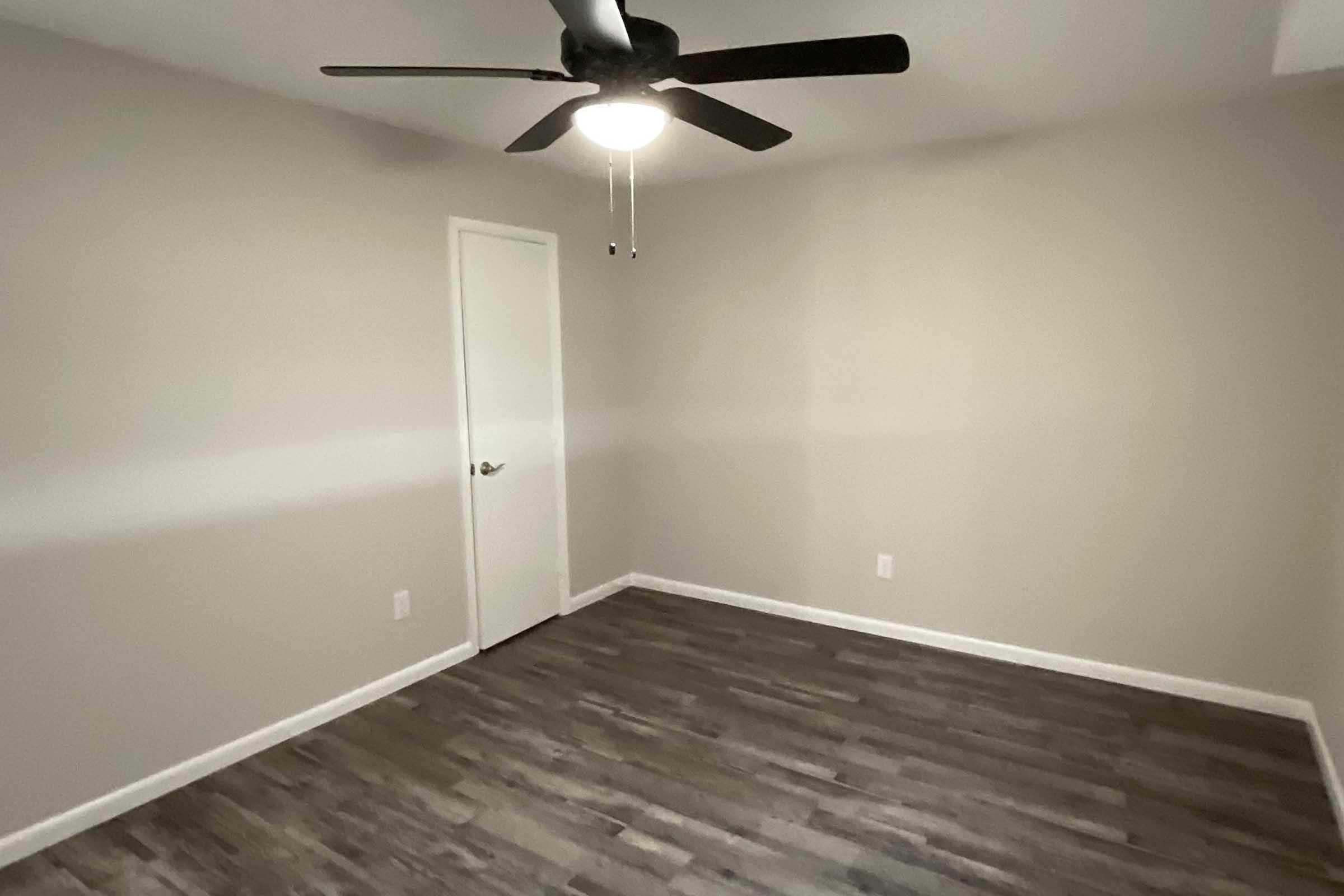 A minimalist room with light gray walls and a ceiling fan. The floor features dark laminate wood flooring. A closed white door is visible on one side, and natural light shines through, creating soft shadows in the space. The room is empty, emphasizing its simplicity and potential for personalization.