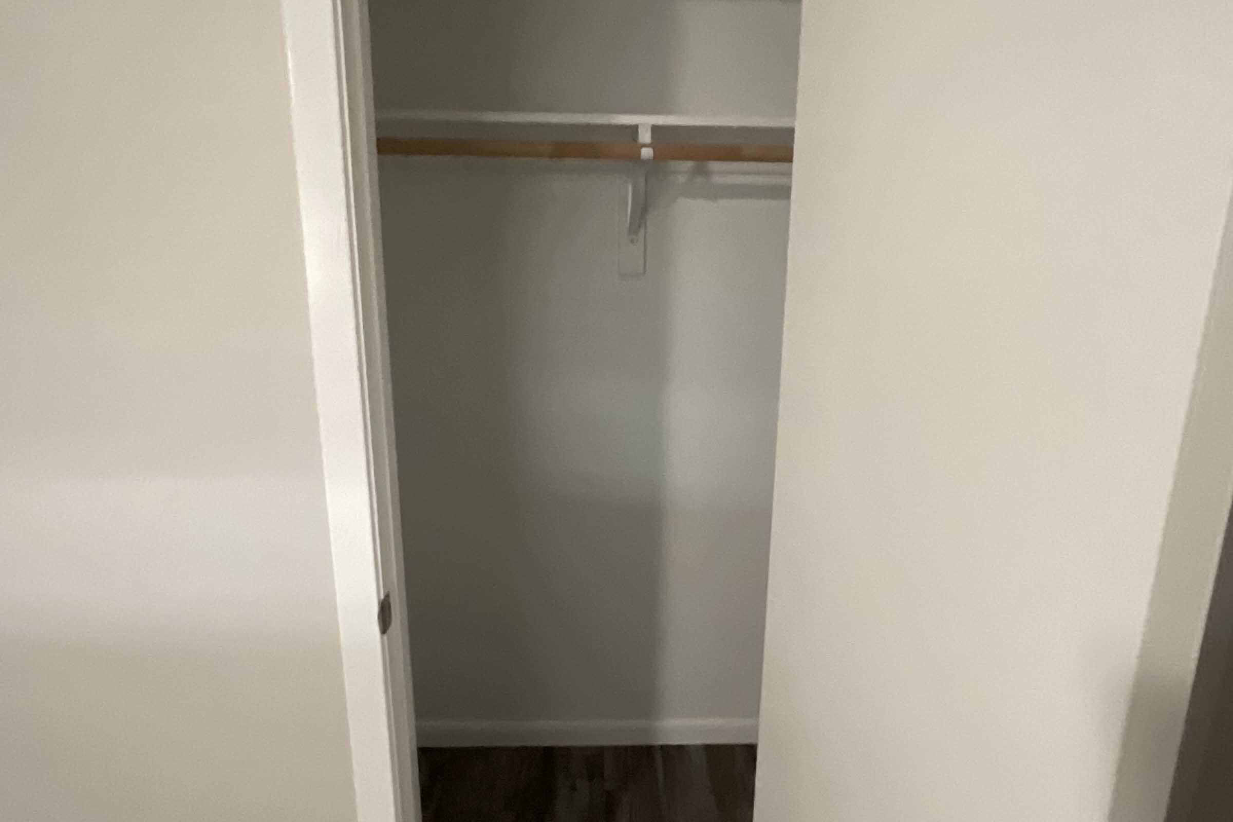 A view of an empty closet with a light-colored wall, a wooden hanging rod, and no items inside. The floor appears to be wooden, and the closet doors are slightly ajar, revealing a spacious, minimalist interior.