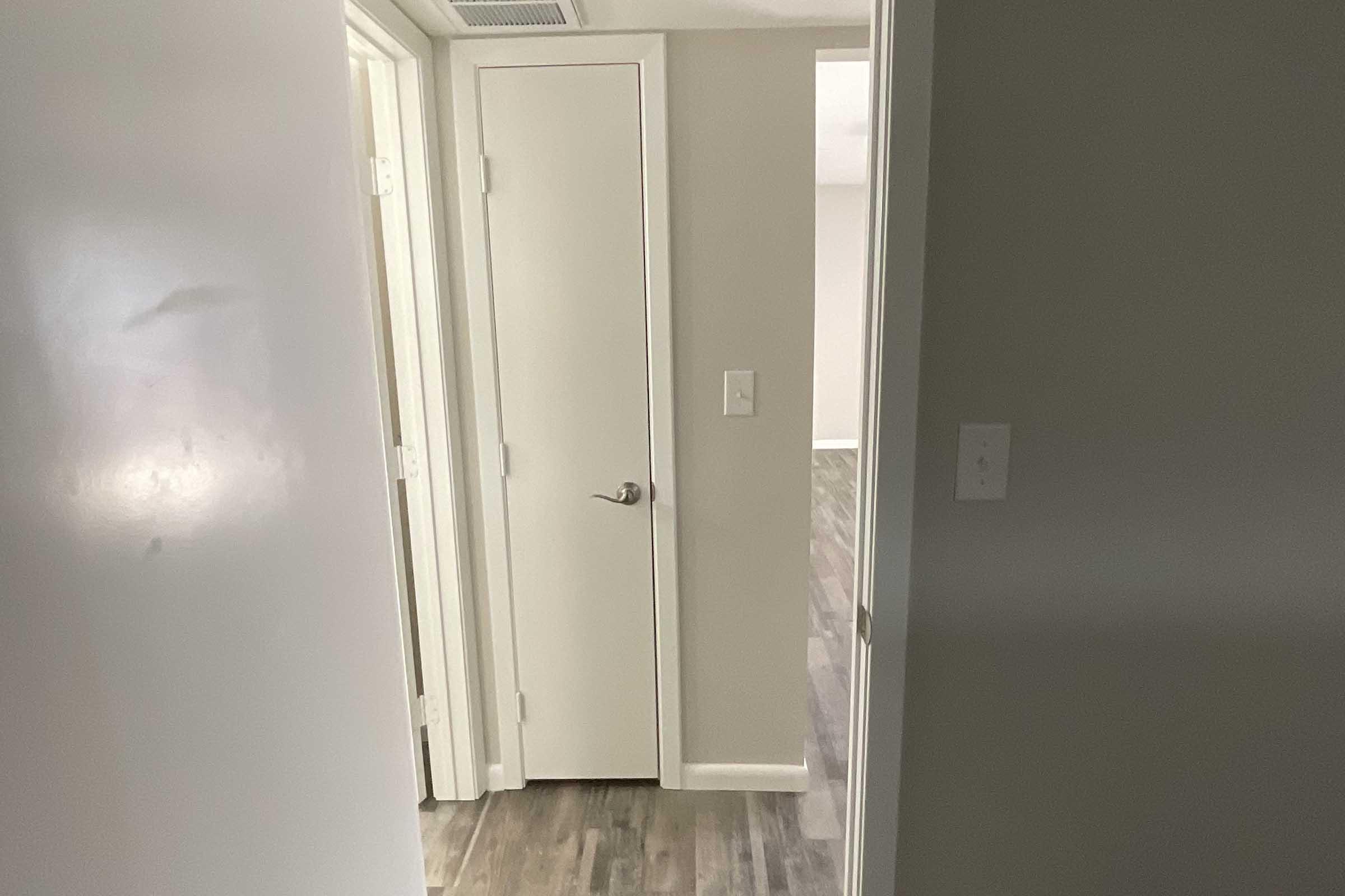 A narrow hallway featuring a white door on the left, leading to another room. The walls are painted in a light neutral color, and the flooring is a wood-like laminate. Another door is visible in the background, suggesting an open layout. Natural light illuminates the space.