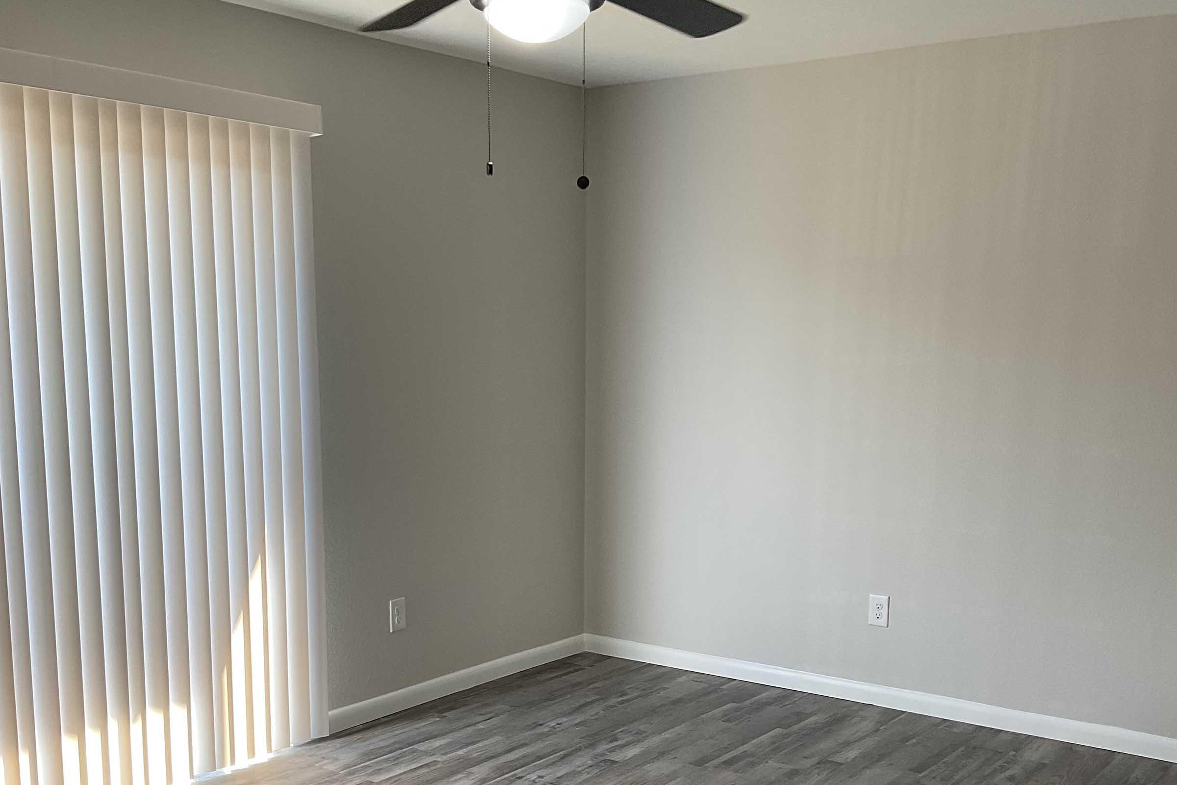 Empty room with light-colored walls and a wooden floor. There is a ceiling fan with a light fixture, and one wall features vertical blinds partially opened to let in light. The overall atmosphere is clean and minimalistic, focusing on space.