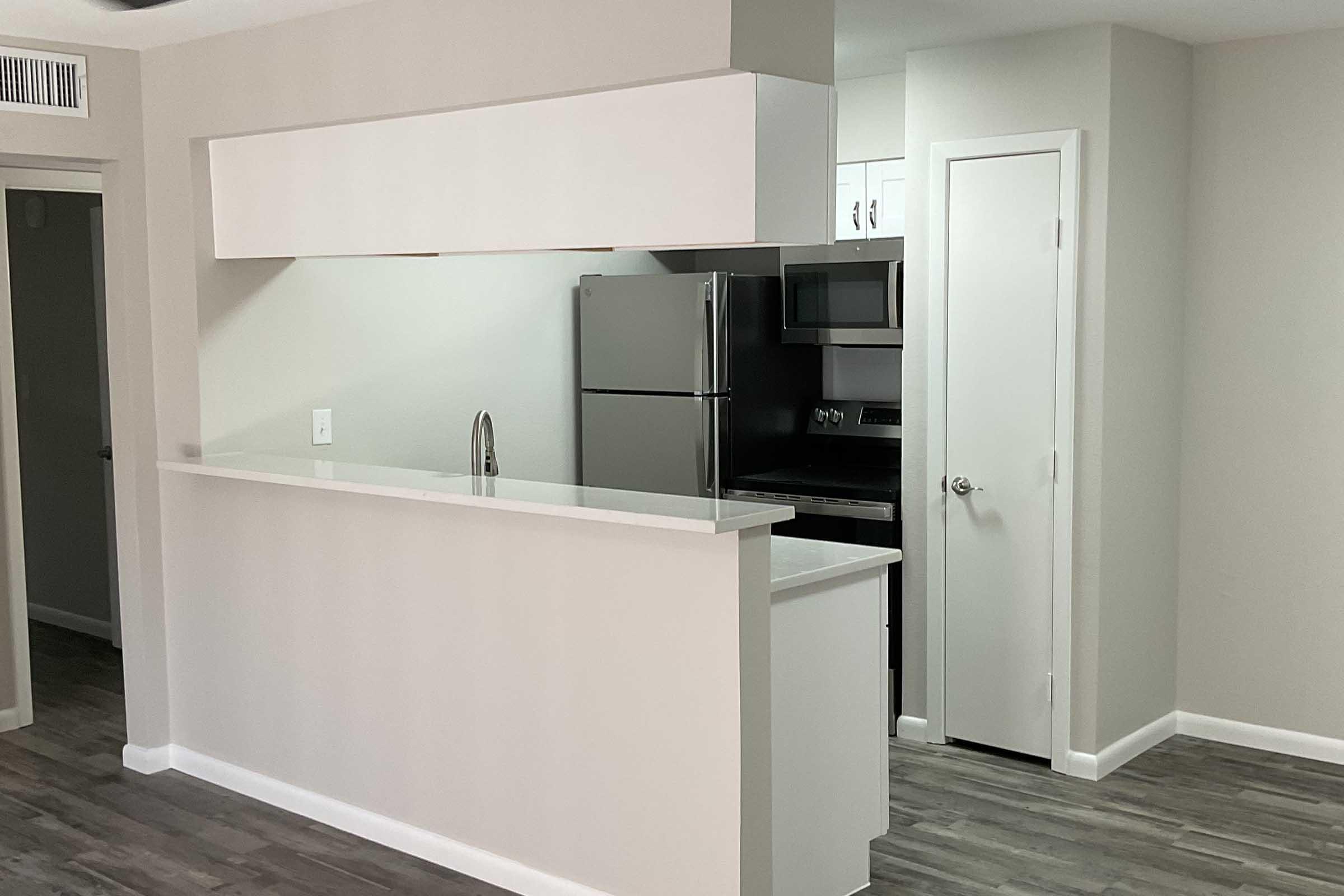 A modern kitchen with a bar-style counter, featuring a stainless steel refrigerator and stove. The cabinetry is minimalist and white, and the room has grey wood flooring. Bright lighting highlights the clean lines and spacious layout. The color palette is neutral, creating a contemporary feel.