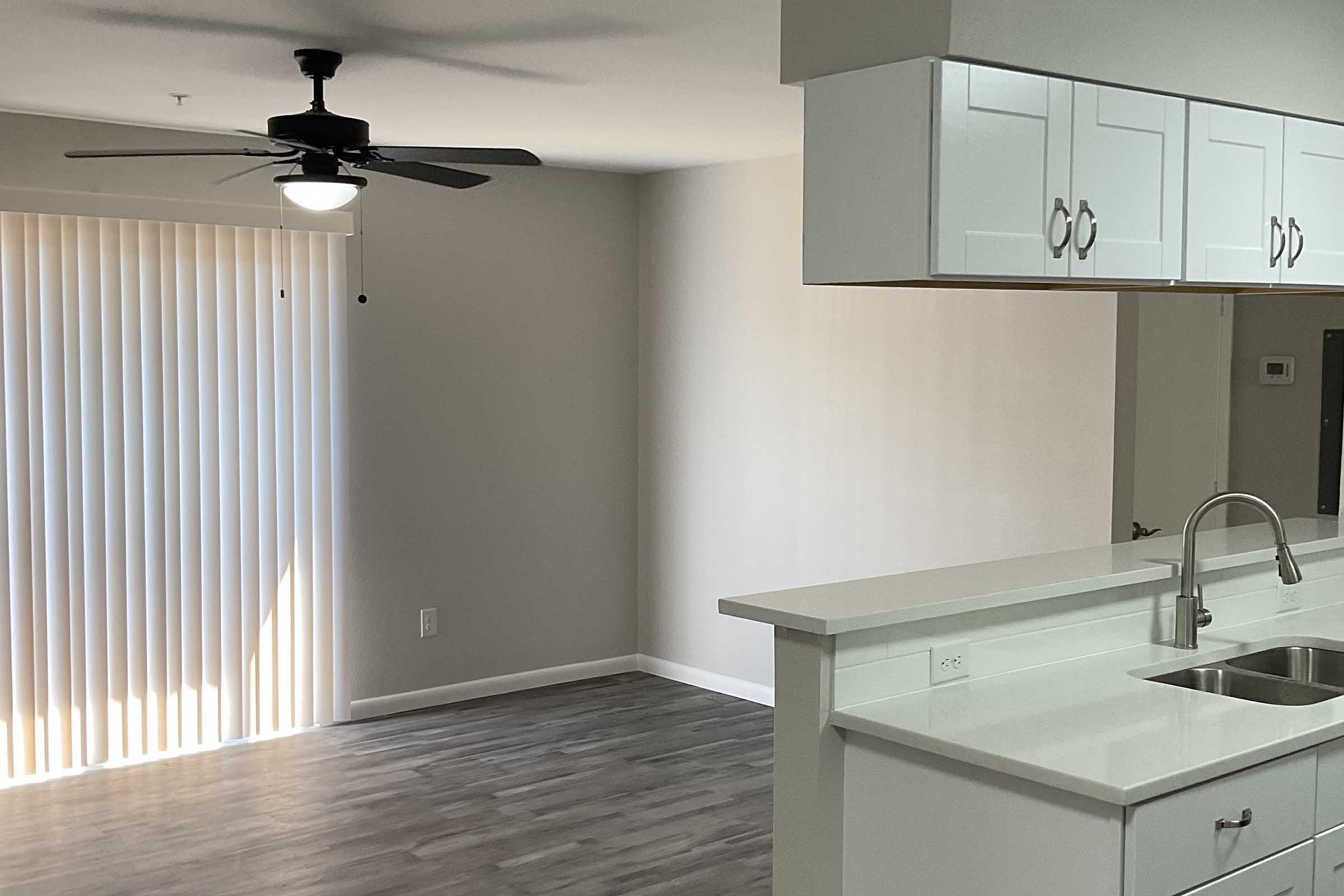 A well-lit modern living space with a ceiling fan, featuring a kitchen area with a white countertop and sink. The room has a large window with vertical blinds, casting soft light onto the light-colored walls and sleek flooring. The open layout creates a spacious feel.