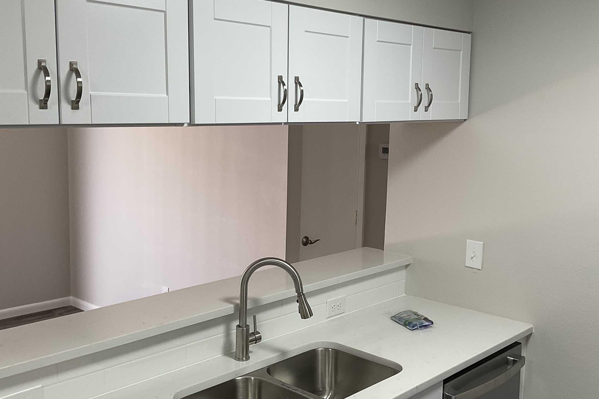 Modern kitchen featuring white cabinetry with silver handles, a stainless steel sink with a polished faucet, and a clean countertop. The background includes a glimpse of a room with light-colored walls and a door, creating a bright and spacious atmosphere.