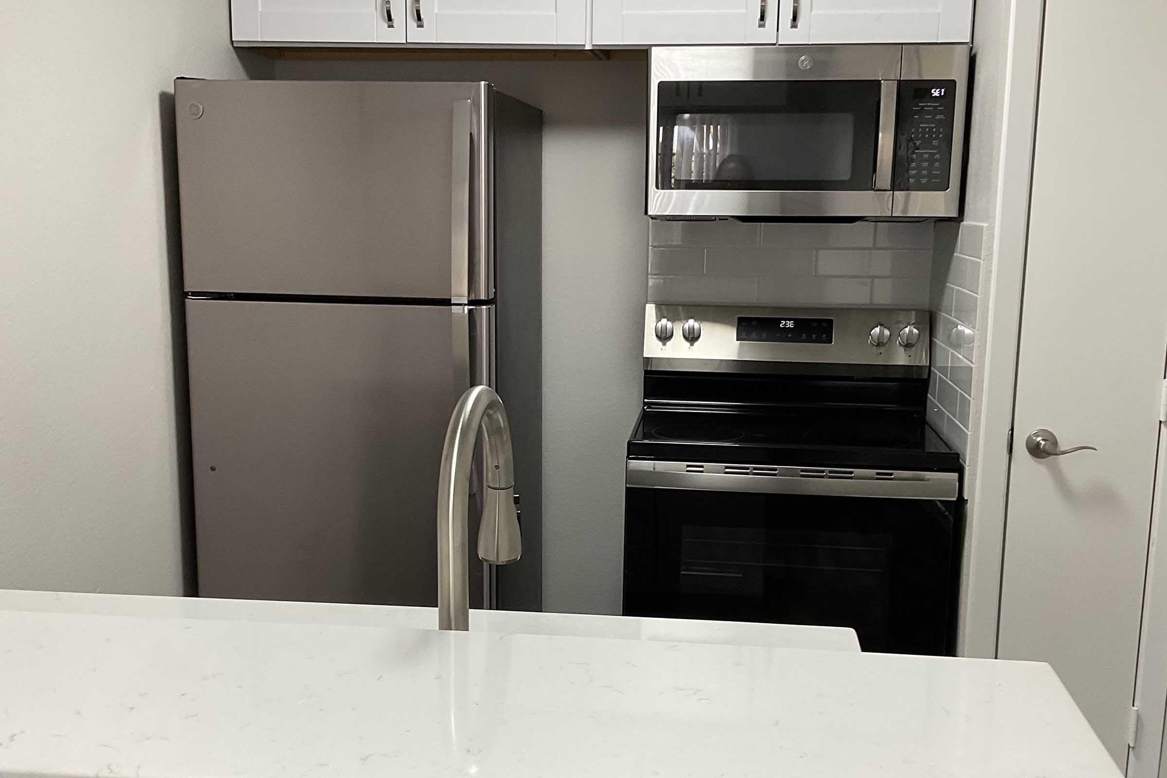 Modern kitchen with stainless steel appliances including a refrigerator, microwave, and oven. There is a sleek faucet above a white countertop, and cabinetry above the appliances. The overall design is contemporary and minimalist, featuring light-colored walls and tile backsplash.