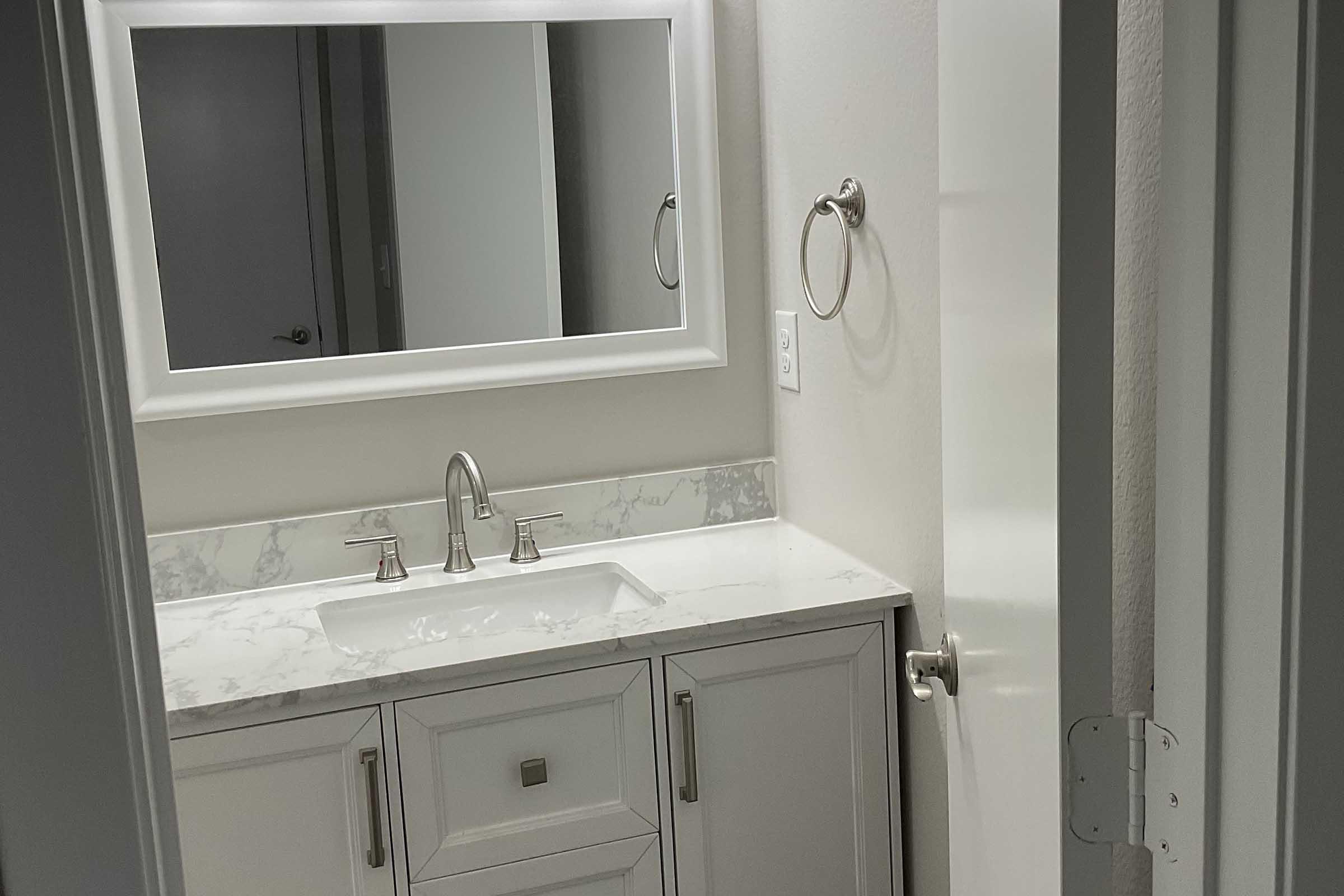 A modern bathroom featuring a white vanity with a marble countertop and sink, a large mirror above the sink, a towel ring on the wall, and a partially open door revealing a glimpse of another room. The space is well-lit and has neutral-colored walls.