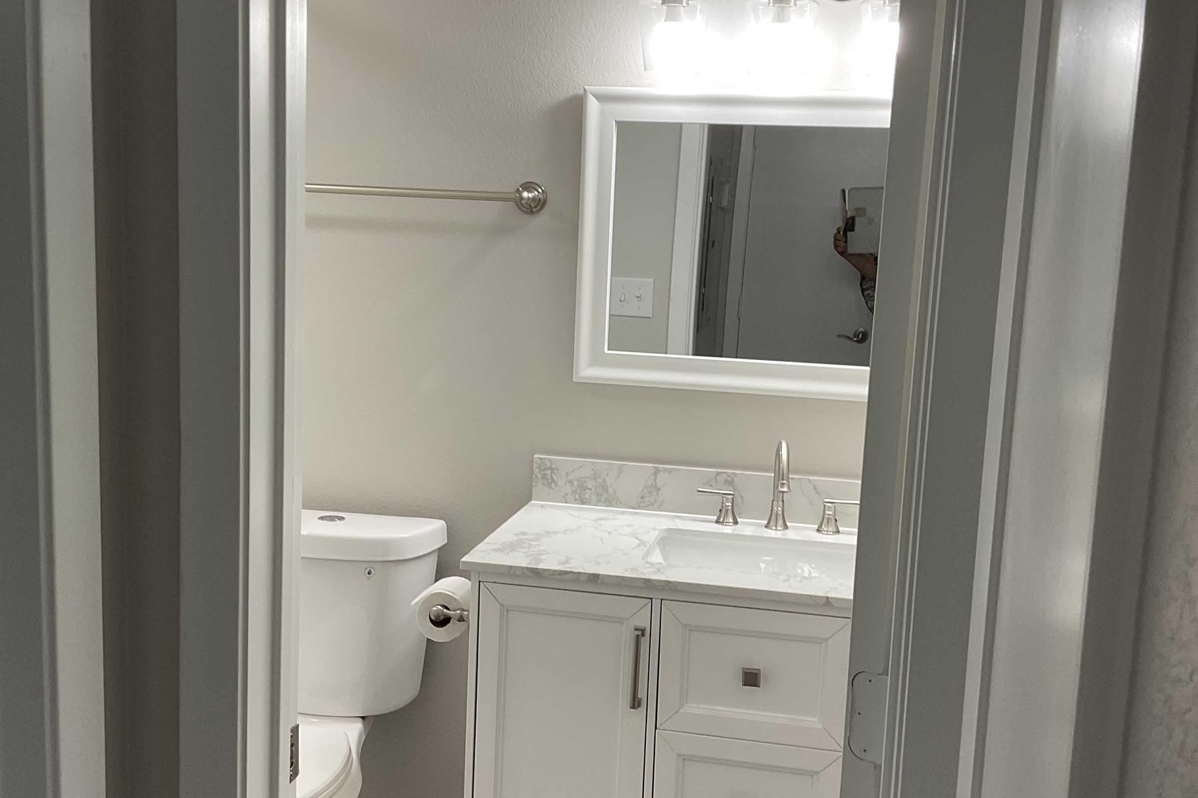 A modern bathroom featuring a white vanity with a marble countertop, a mirror above the sink, a towel bar on the wall, and a toilet in the corner. The walls are painted in a light color, and there is warm lighting from above, creating a bright and inviting atmosphere.