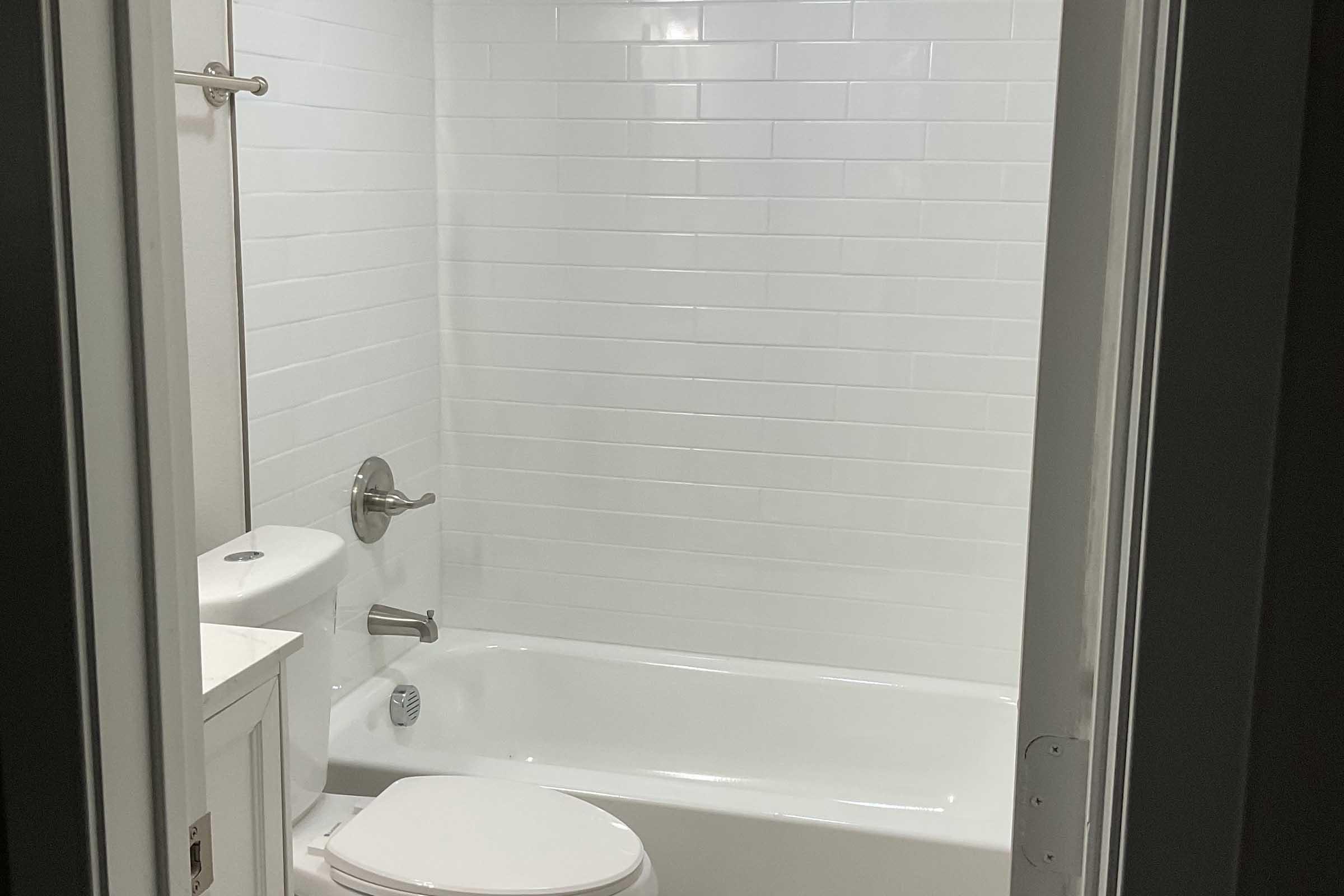 A clean, modern bathroom featuring a white tiled wall, a bathtub with a shower, and a white toilet. The bathroom is well-lit, creating a bright, spacious appearance. The doorframe is visible, indicating the entrance to the bathroom.