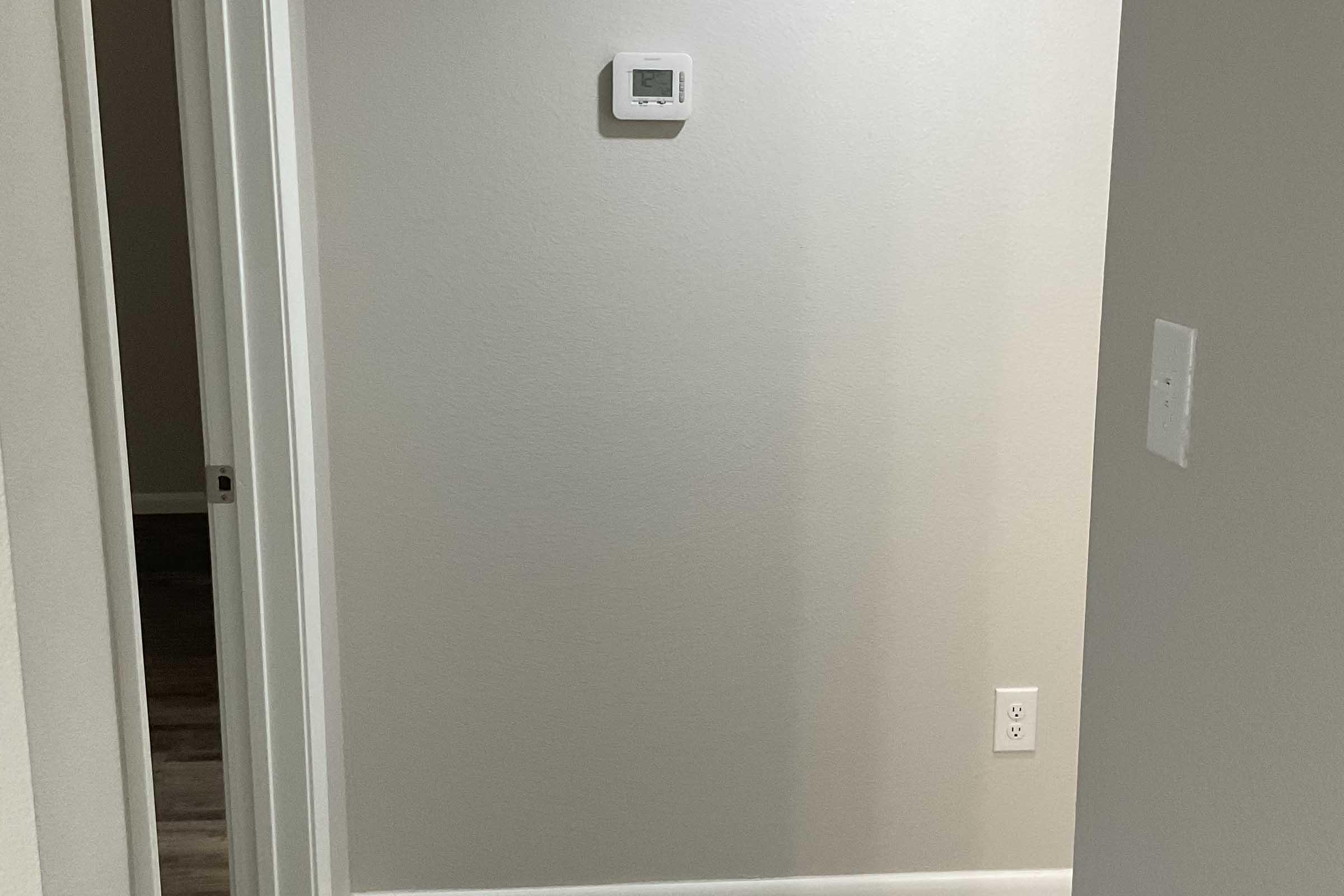 A view of a light-colored interior wall with a thermostat mounted on it. On the right, a white electrical outlet is visible. The image captures an empty hallway leading to another room, with soft lighting and a simple, modern aesthetic.