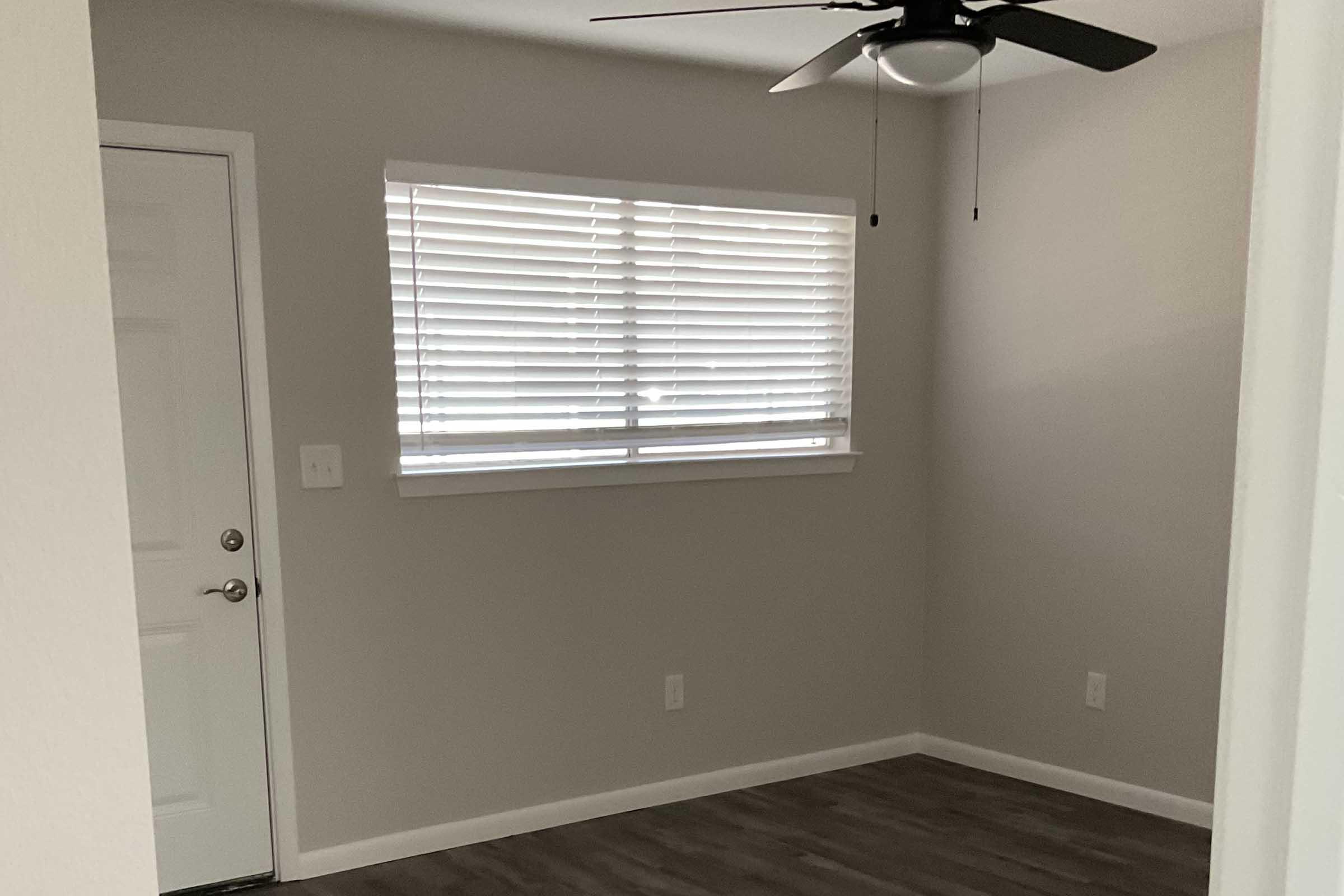 A small, empty room with light gray walls and a ceiling fan. There is a closed white door on the left and a window with white blinds that allows natural light to enter. The floor is wood laminate, creating a clean and modern appearance.