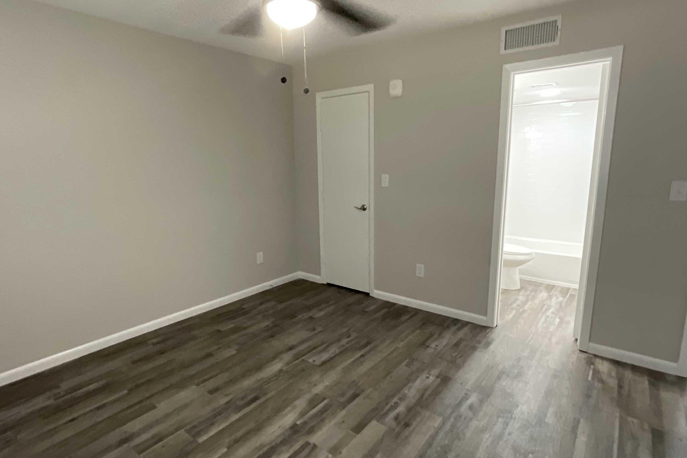 A neutral-colored room with gray walls and a wooden floor. A ceiling fan is visible, and there is a closed white door leading to another room. In the background, a bathroom is partially visible, featuring a tub and toilet. The space is bright and uncluttered, suitable for various uses.