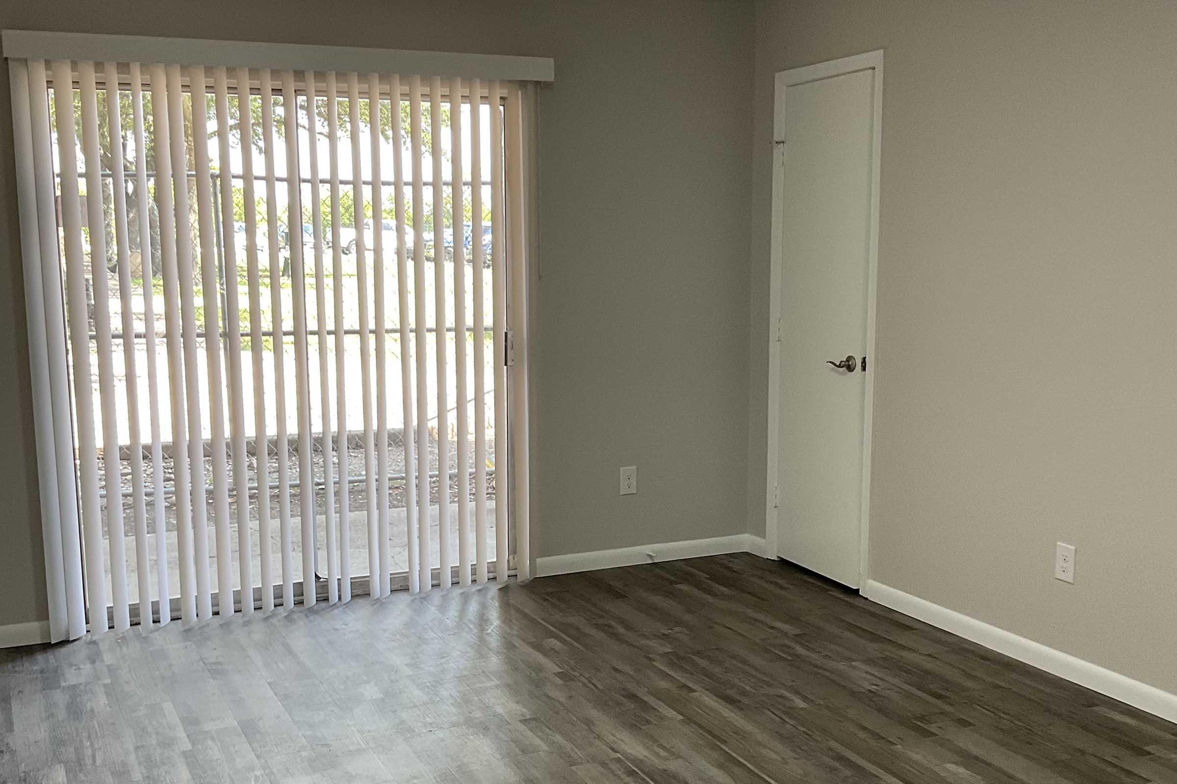 A minimalistic interior room featuring a light gray wall and a wooden floor. A door is visible on the right, and next to it, vertical blinds cover a large window, allowing natural light to enter the space. The room appears empty and ready for furnishing.