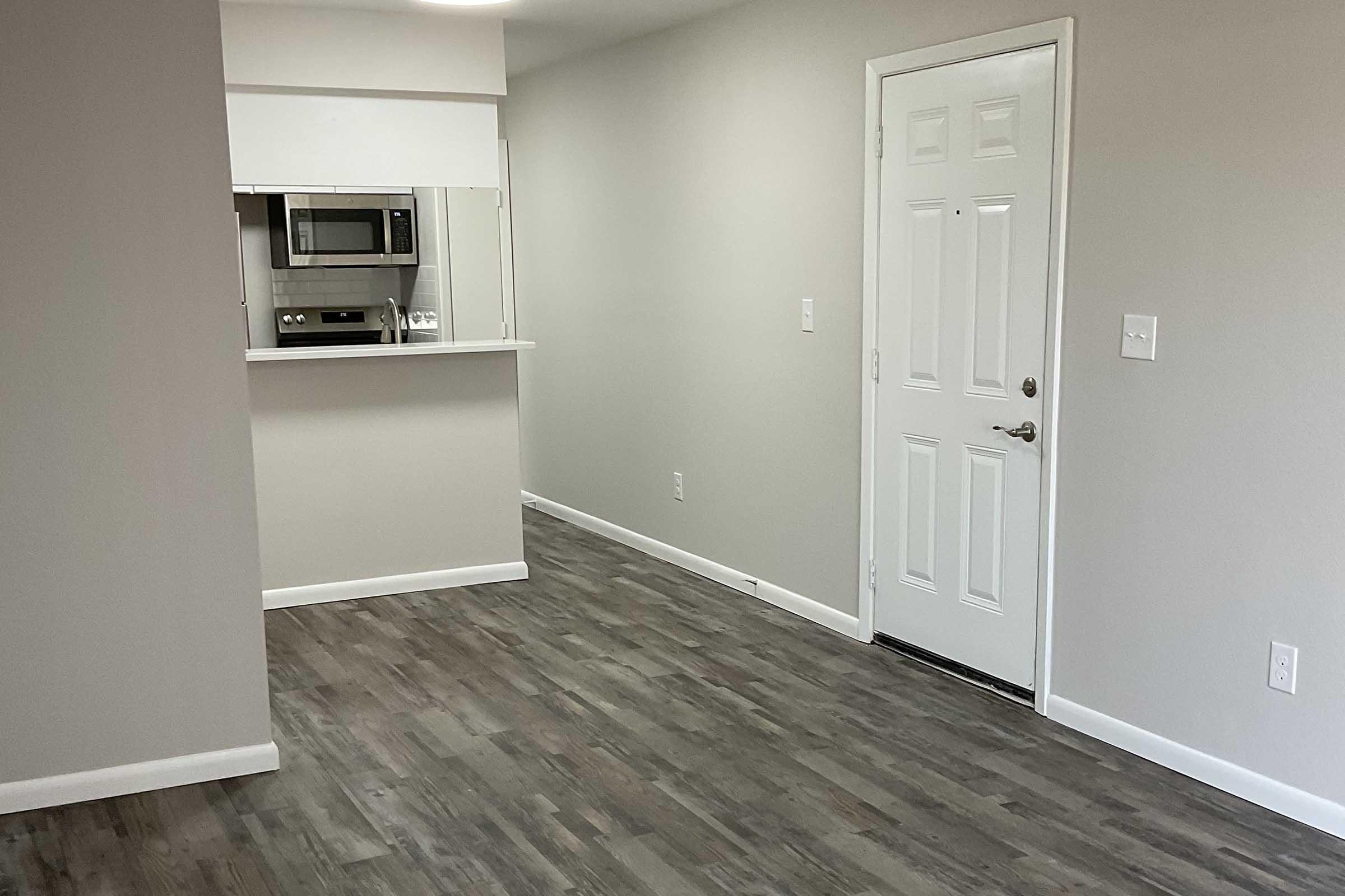 A view of a modern, empty living space featuring light gray walls, a wooden floor, and a white door. A small kitchen area is visible in the background, with a microwave and a counter. The room is well-lit, creating a spacious and contemporary atmosphere.