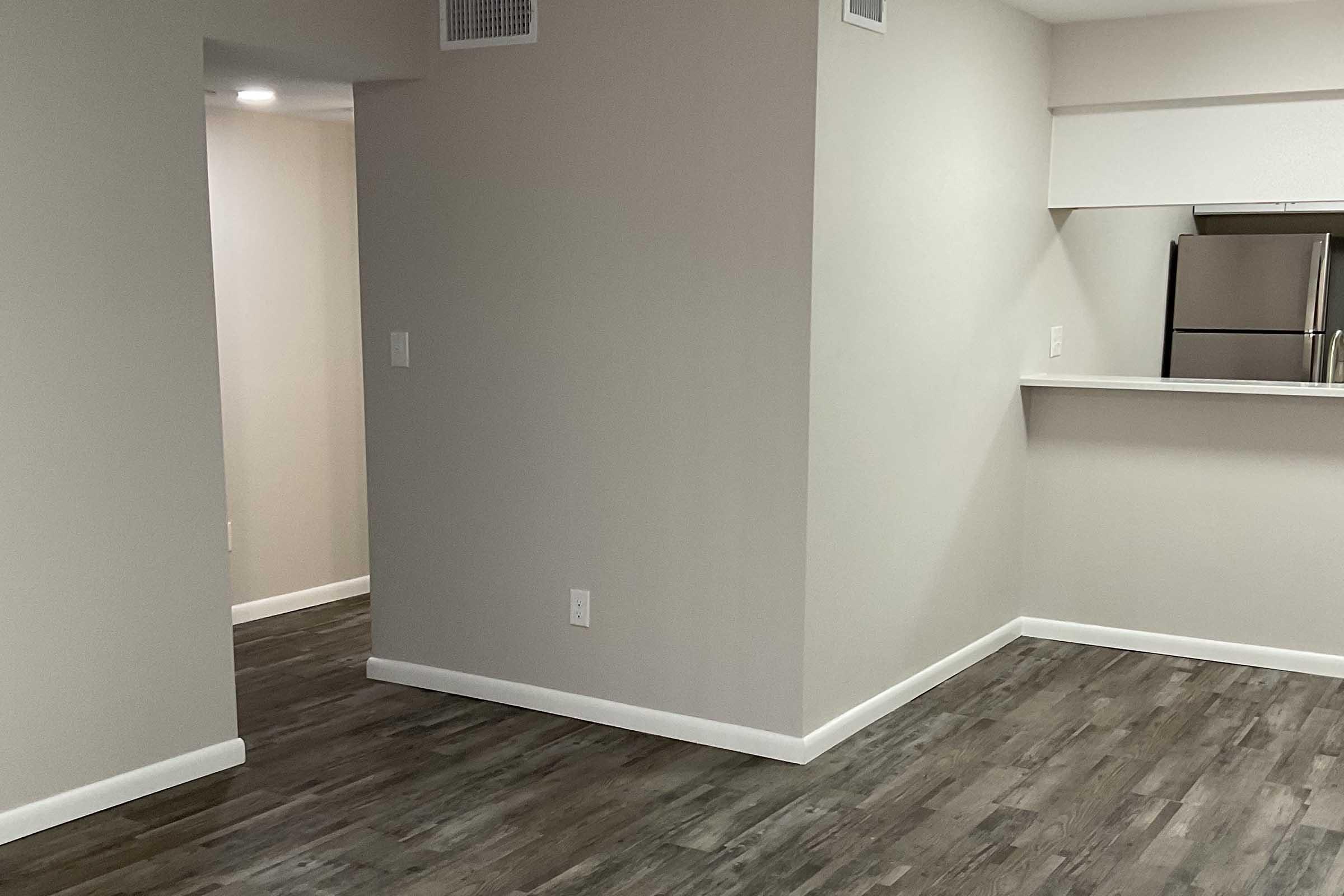 Spacious interior of an apartment with light gray walls and wood-like flooring. The layout includes an open living area leading to a kitchen with stainless steel appliances. A doorway on the left leads to another room, enhancing the sense of openness and connectivity in the space. Natural light illuminates the area.