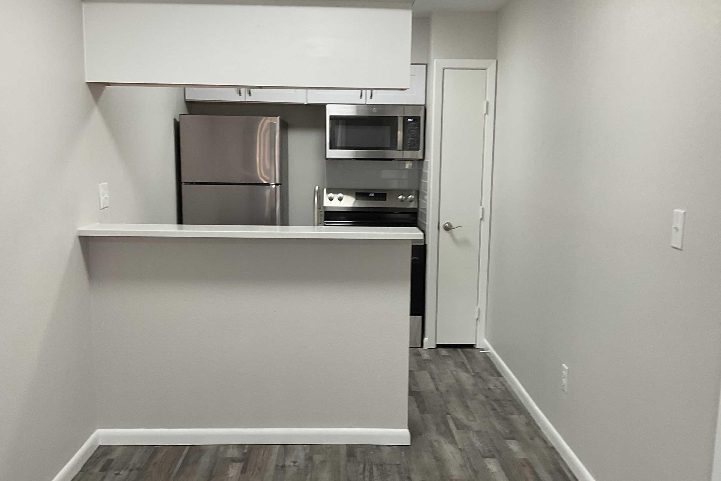 A modern kitchen space featuring a stainless steel refrigerator, microwave, and oven. The kitchen includes a countertop with a small bar area, light-colored walls, and laminate flooring. The design is sleek and functional, showcasing an open layout.