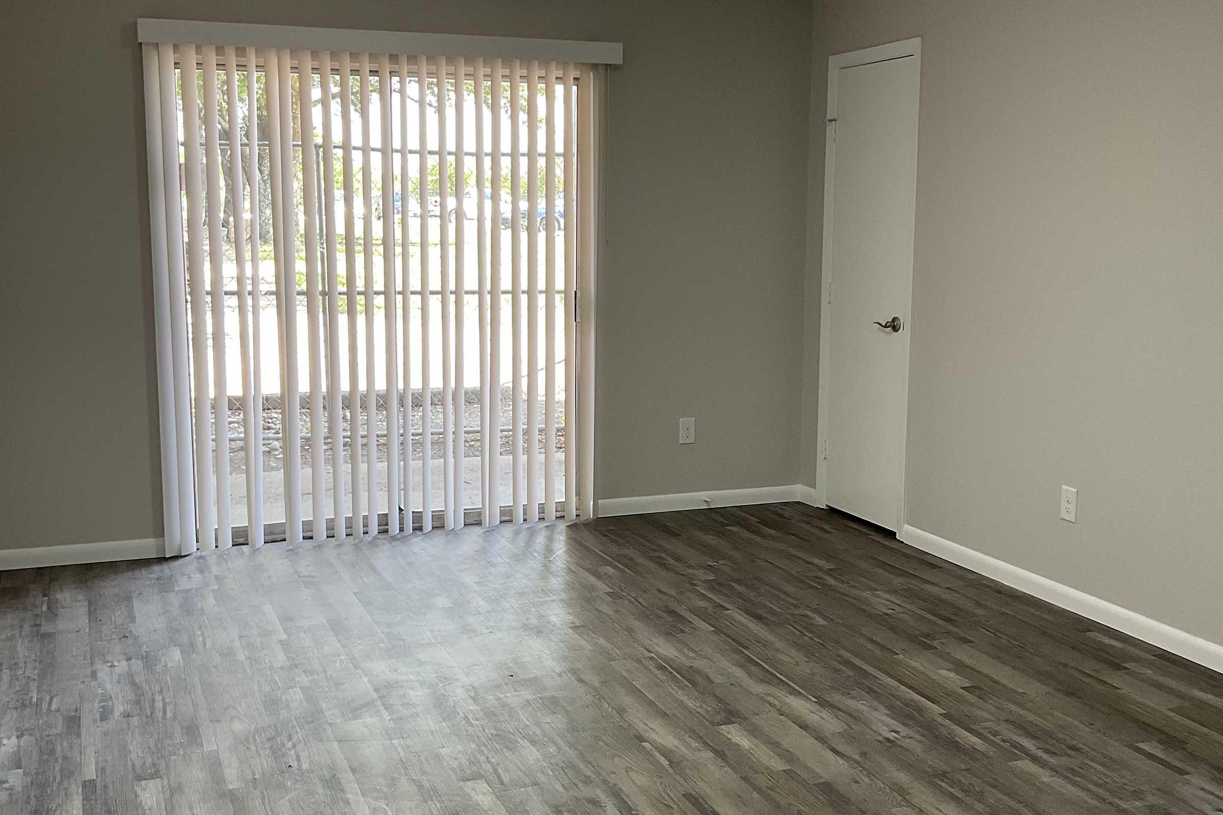 An empty room with light brown flooring, soft gray walls, and a large window with vertical blinds partially open, allowing natural light to enter. A white door is visible on the right side of the image. The space appears bright and ready for furniture arrangement.