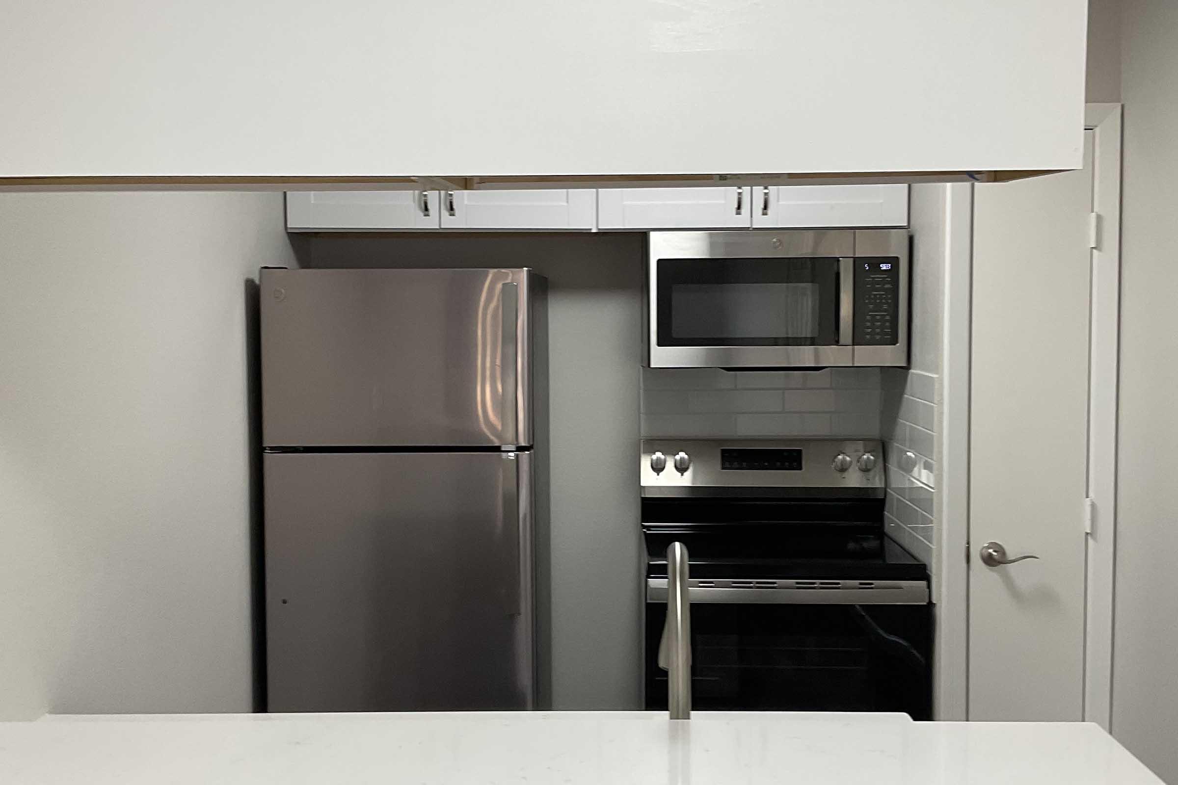Modern kitchen featuring a stainless steel refrigerator, microwave, and black oven with a smooth cooktop. White cabinetry above and a clean, minimalist countertop. Light-colored walls and a door visible in the background.