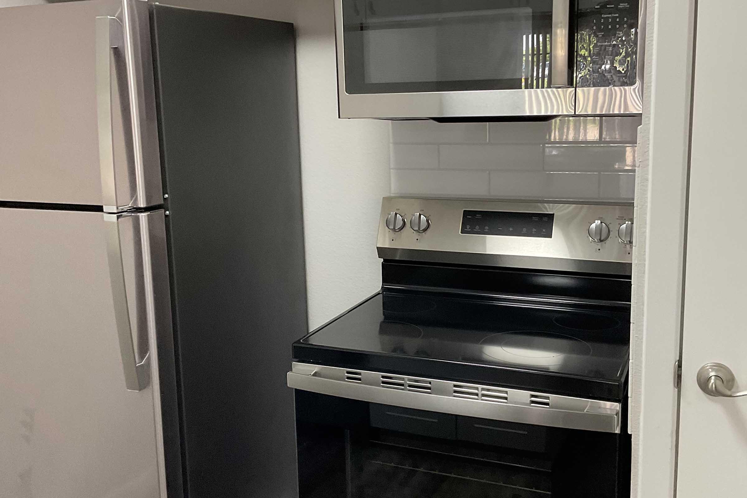 A modern kitchen featuring a stainless steel refrigerator on the left, an electric stove with an oven beneath it in the center, and a microwave mounted above. The walls are white, with a tiled backsplash behind the stove, and the floor has dark tiles.