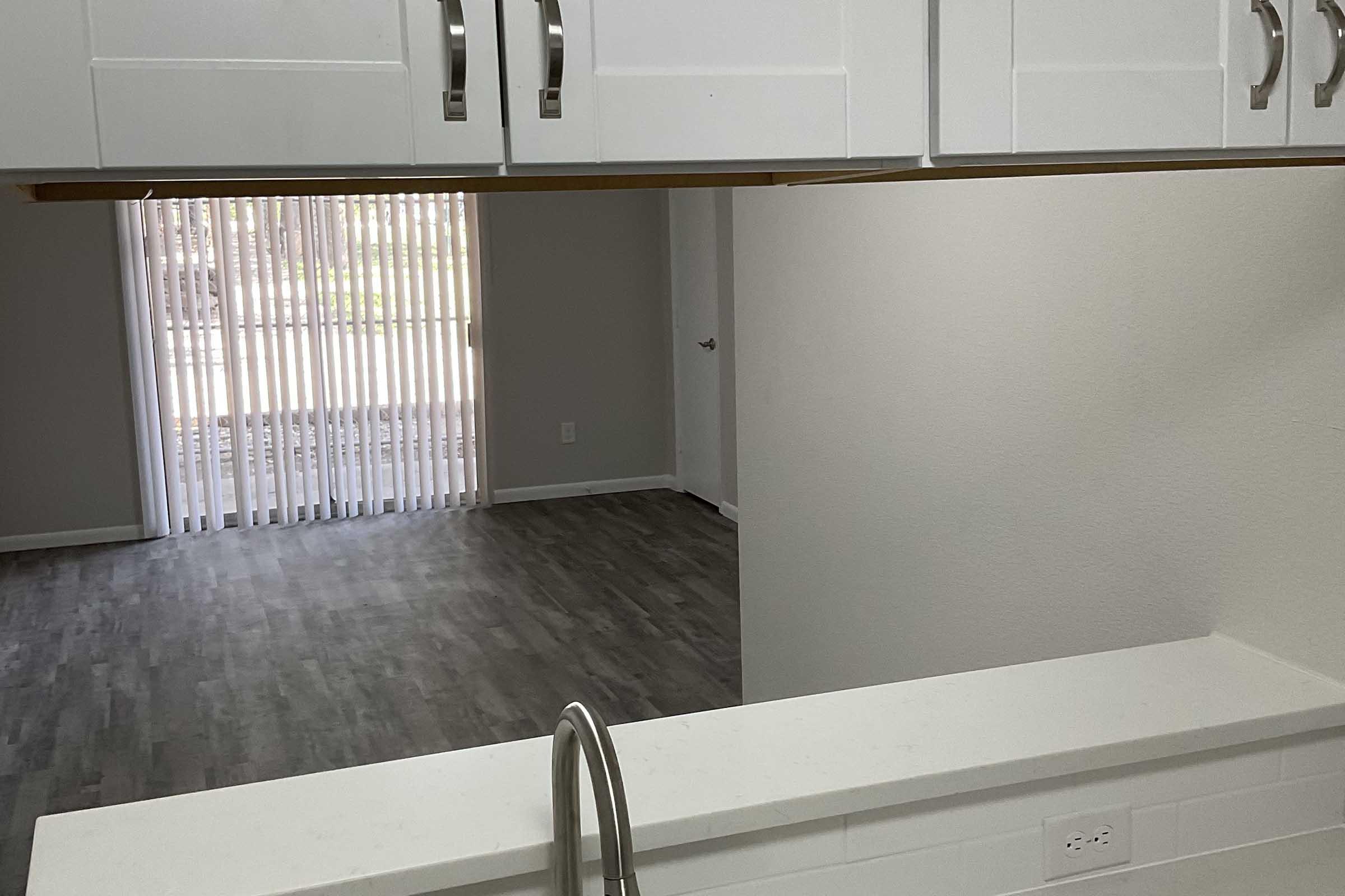 View from a kitchen counter into an open living space. The area has light gray walls, a window with vertical blinds, and a doorway visible in the background. Flooring appears to be laminate, resembling wood. The kitchen features white cabinetry and a modern sink.