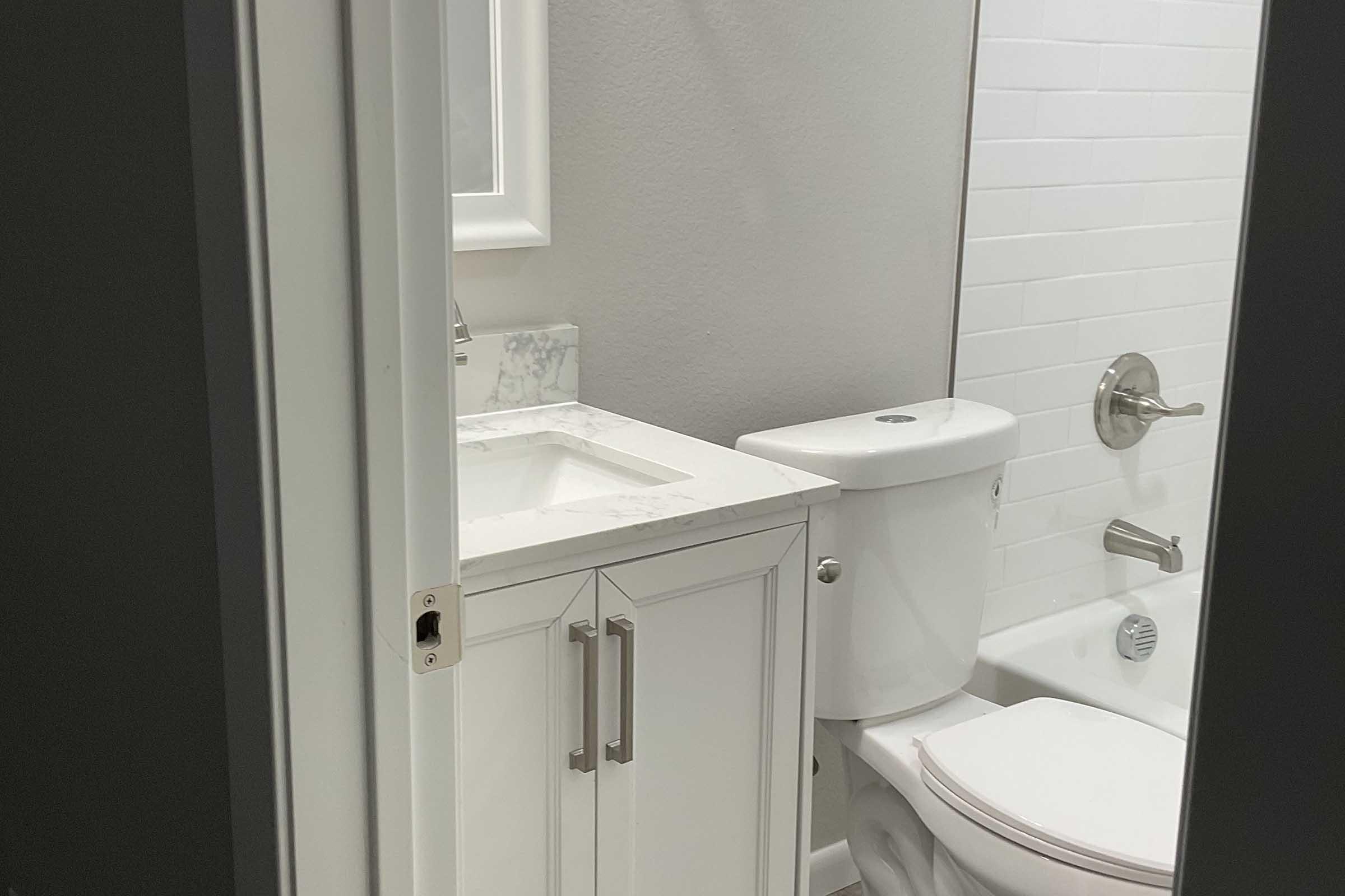 A small bathroom featuring a white toilet, a sink with a marble countertop, and a modern shower with white tiles. The space is well-lit, with a mirror above the sink, and the walls are painted in a soft gray color. A door partially frames the view into the bathroom.