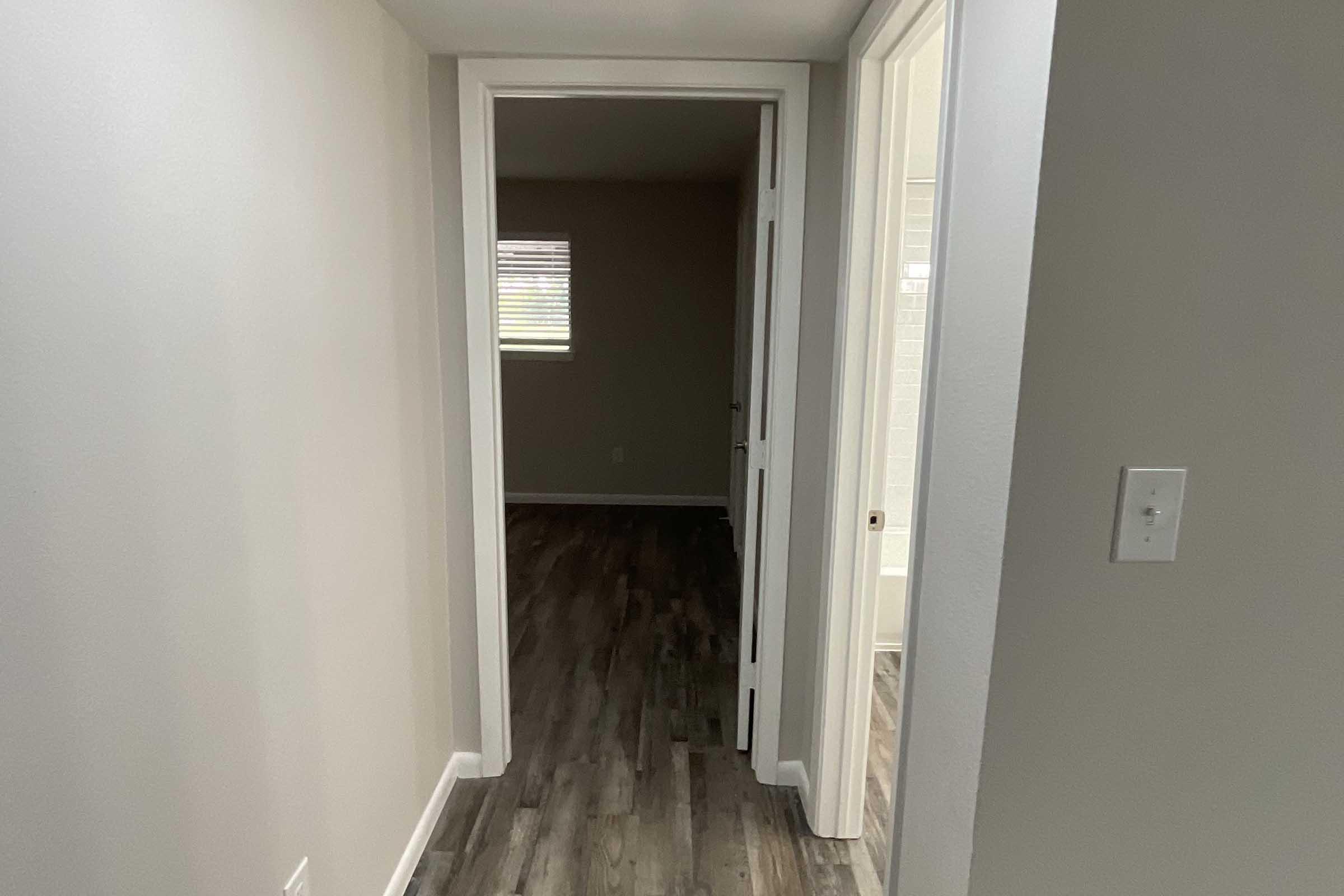 A narrow hallway with light-colored walls and a wooden floor. A door is partially visible on the right, leading to a small room with a window. The hallway is well-lit, and there are no decorations. The overall atmosphere is clean and minimalistic.