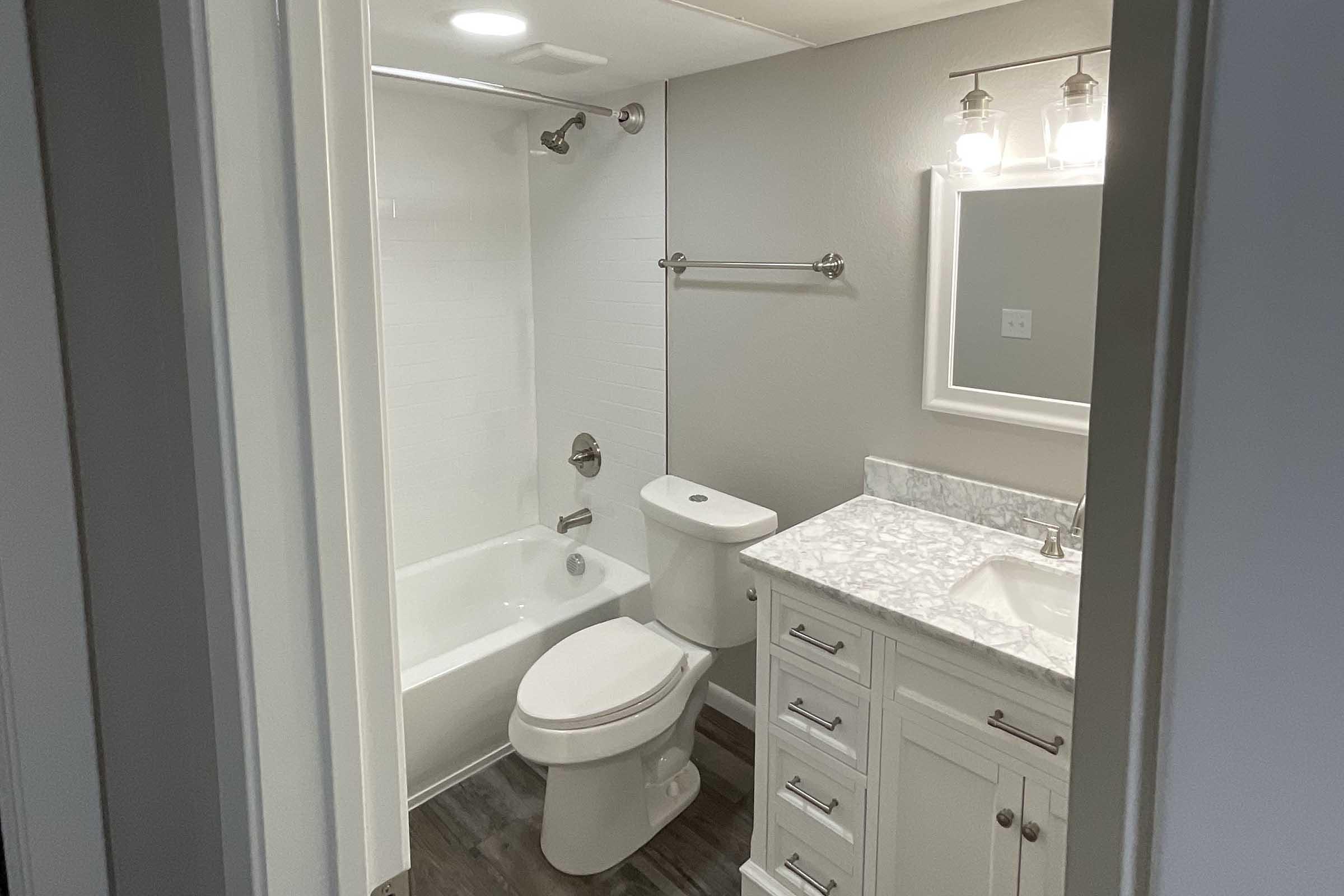 A modern bathroom featuring a white bathtub/shower combo, a toilet, and a vanity with a marble countertop and sink. The walls are painted gray, and there are three pendant lights above the mirror. The floor has a dark wood finish, and the room is well-lit.