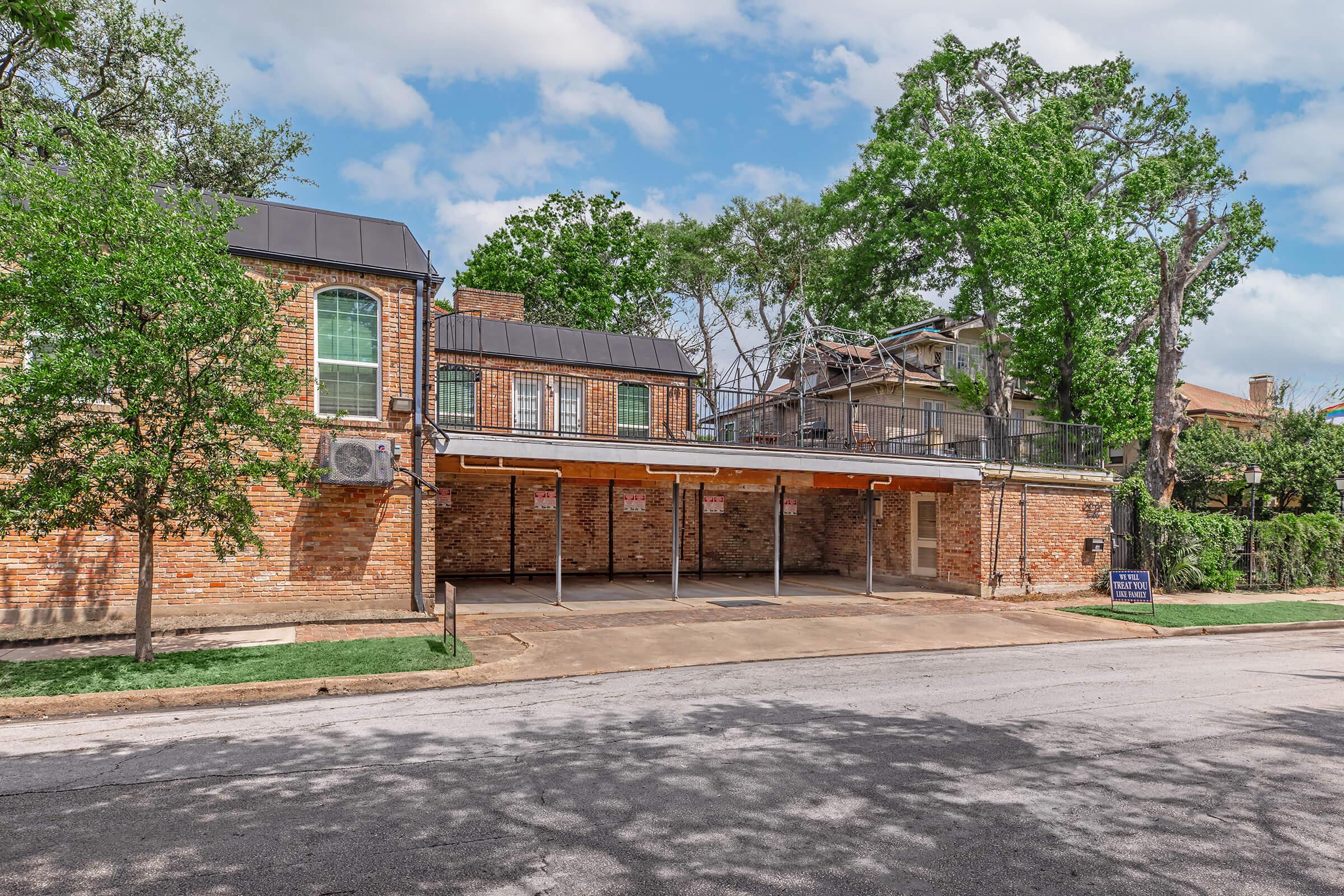A brick building with a covered parking area in front, surrounded by trees and greenery. The structure features large windows, a black roof, and an upper balcony. A sign is visible near the parking area. The scene is set on a quiet street under a cloudy sky.