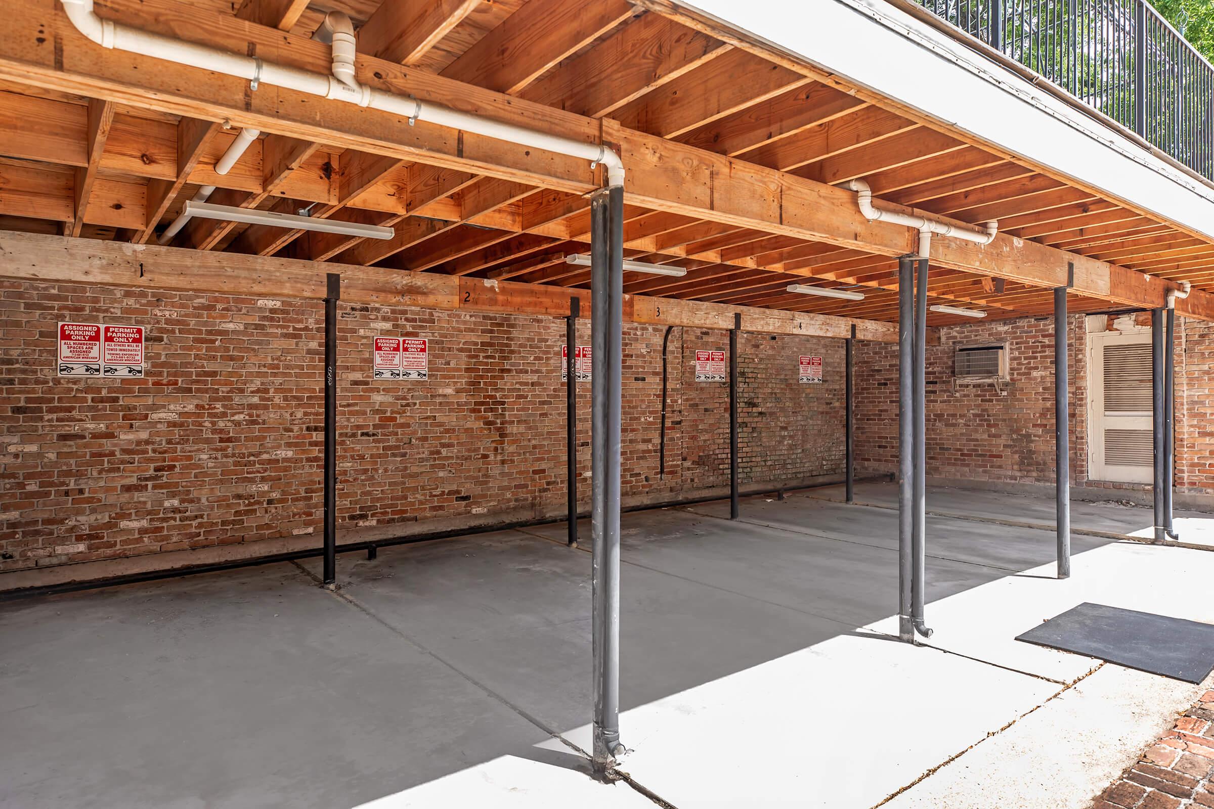 A covered parking area with exposed concrete flooring and brick walls. The structure has wooden beams and metal poles supporting the ceiling. Several warning signs are affixed to the walls, and natural light illuminates the space.