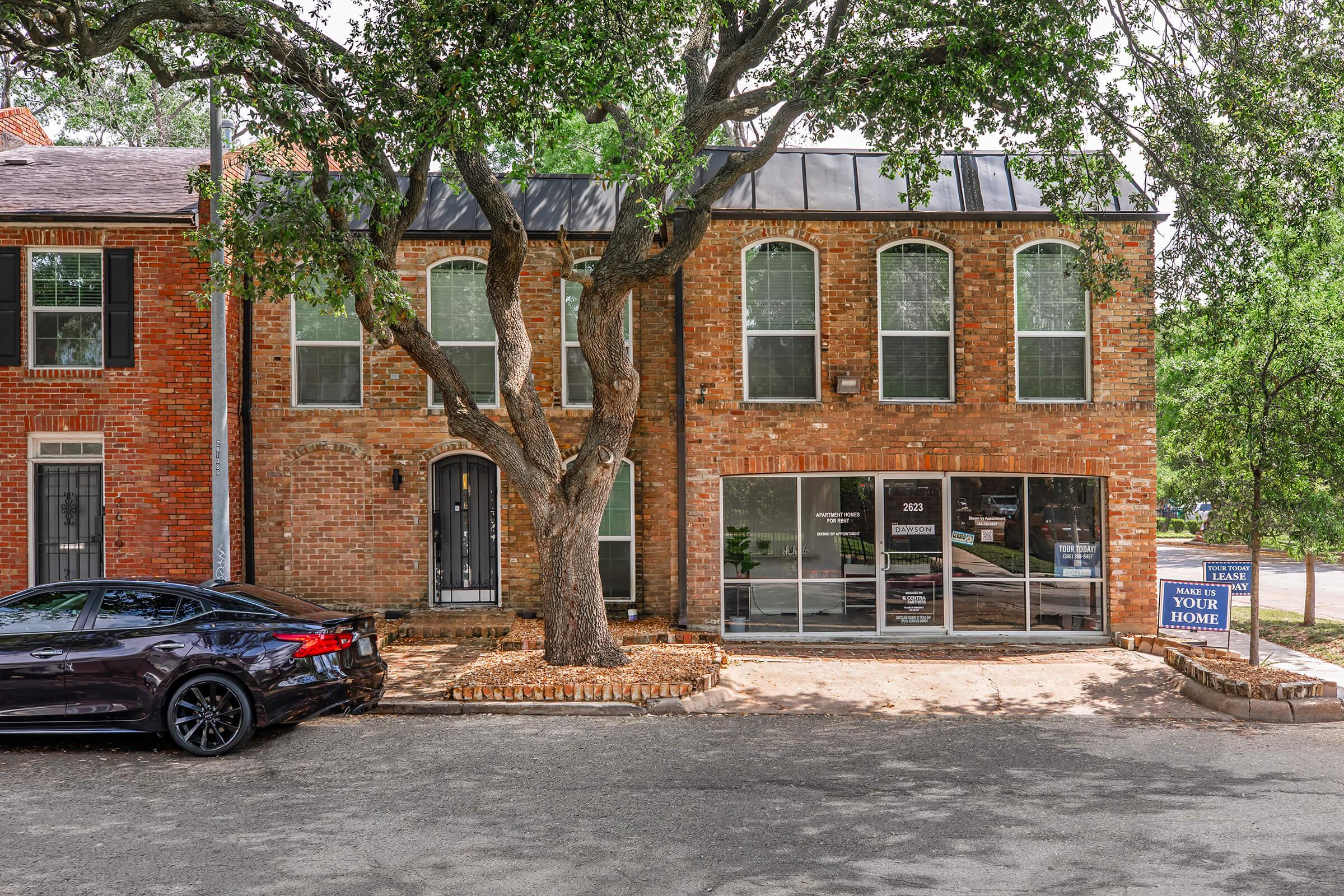 A brick duplex with large windows, featuring a tree in the front yard. One side shows a "For Sale" sign, while the other side appears to be an occupied residence. A black car is parked in front, and the surrounding area includes a landscaped yard and a residential street.