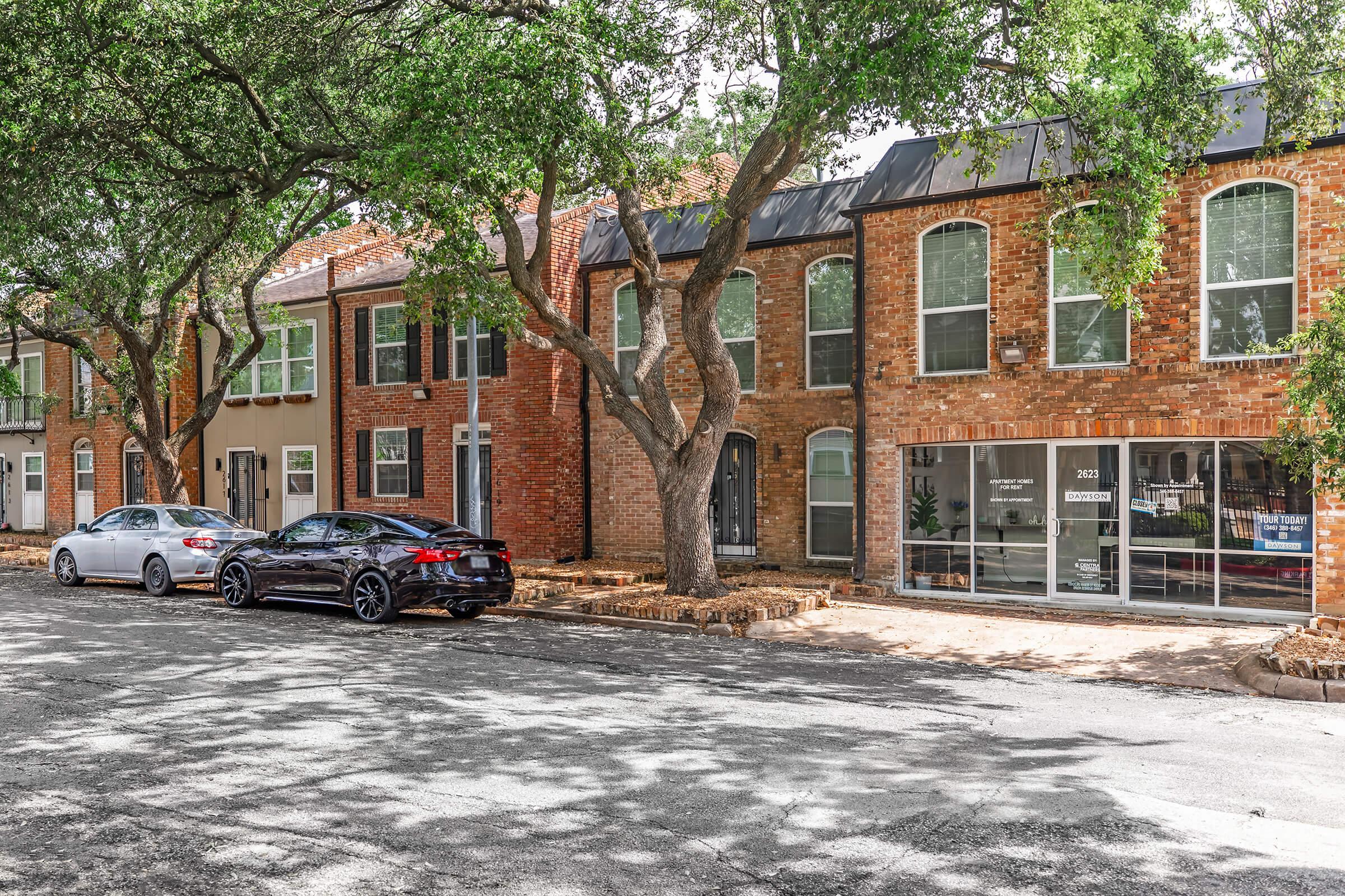 A street view featuring a row of brick townhouses with large windows, shaded by trees. Two cars are parked alongside the curb; one is silver, and the other is black. The scene captures a quiet residential area with a blend of homes and small business storefronts visible through the windows.