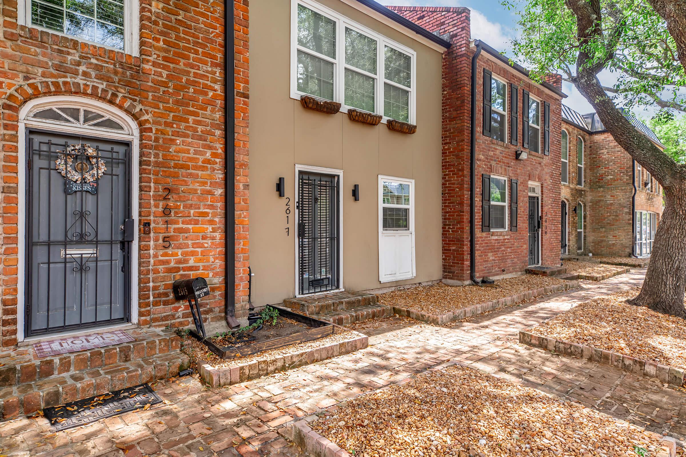A row of charming brick and stucco townhouses with various styles of doors and windows. The pathway is lined with small gravel, and there are trees providing shade to the area. Each unit has a unique character, showcasing a mix of historical and modern architectural elements.