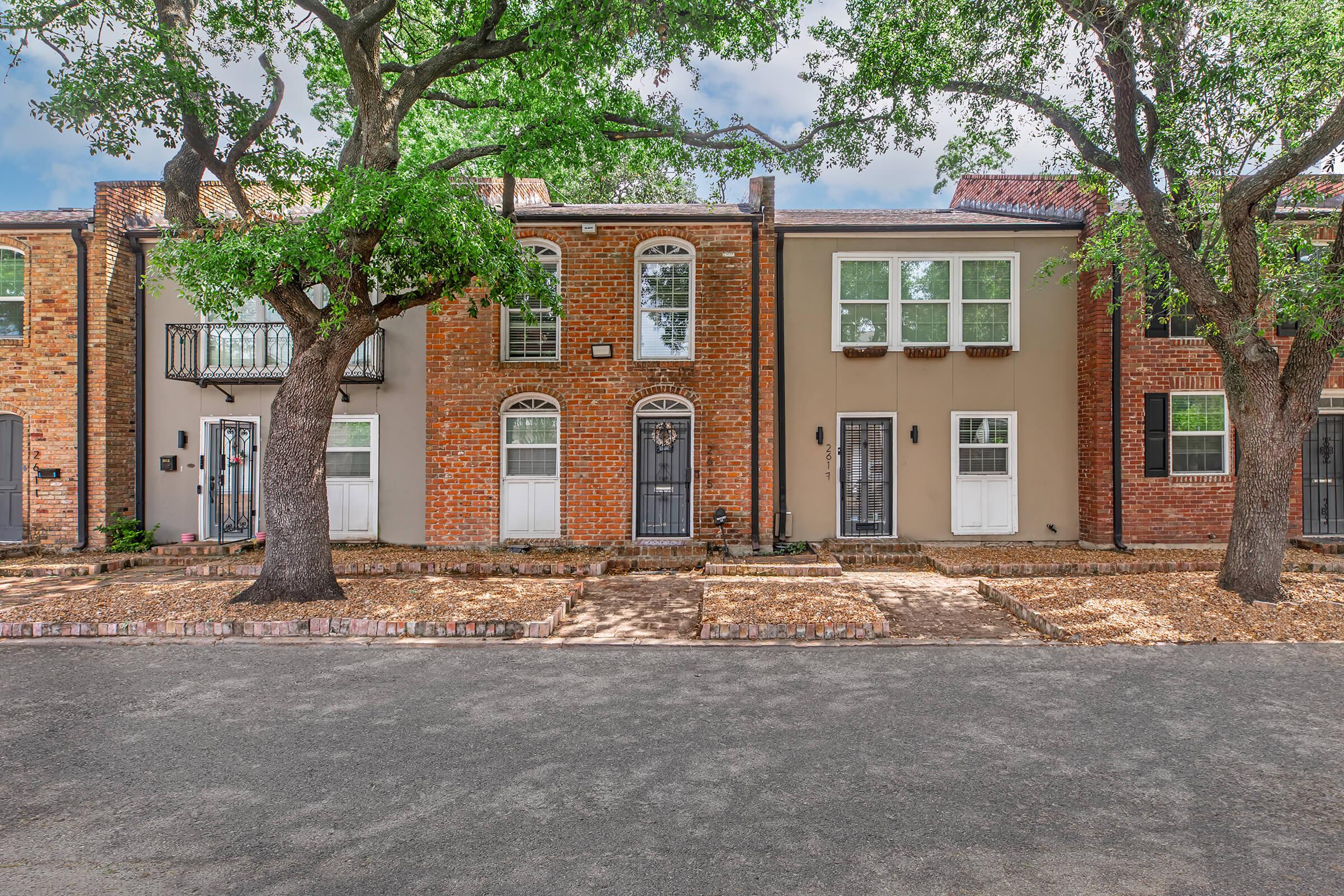 A row of three residential houses with varying brick and stucco facades, flanked by green trees. The first house has a black wrought-iron railing, the second features a mix of light colors and a decorative front window, and the third is painted beige with a white door. The street is paved with pebbles, and there is a clear blue sky above.