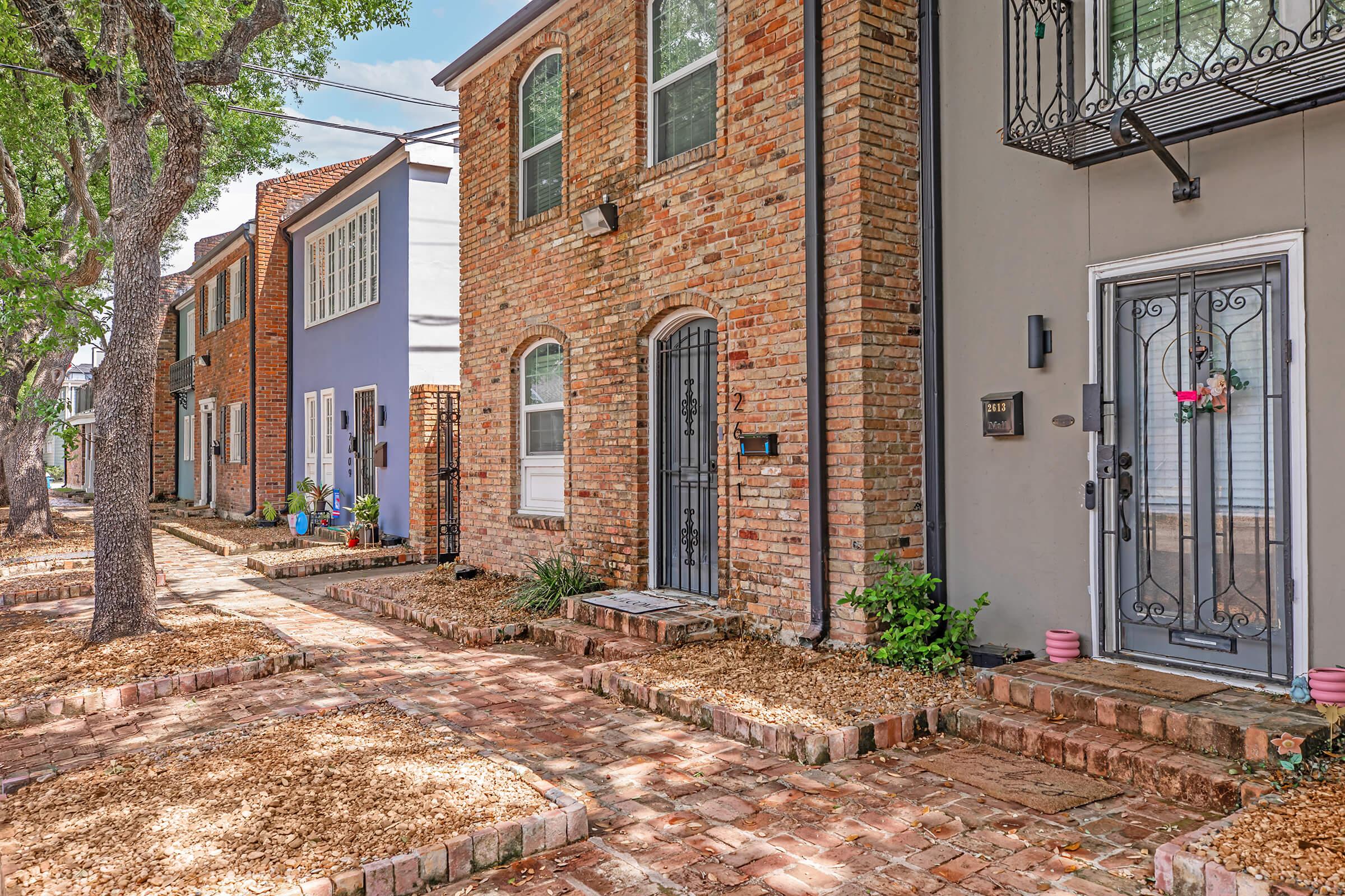 A quaint street view showcasing a row of charming brick and colored houses. Each house features distinct architectural details, such as wrought iron balconies and decorative doors. The pathway is lined with small plants and gravel, adding to the inviting atmosphere of the neighborhood.