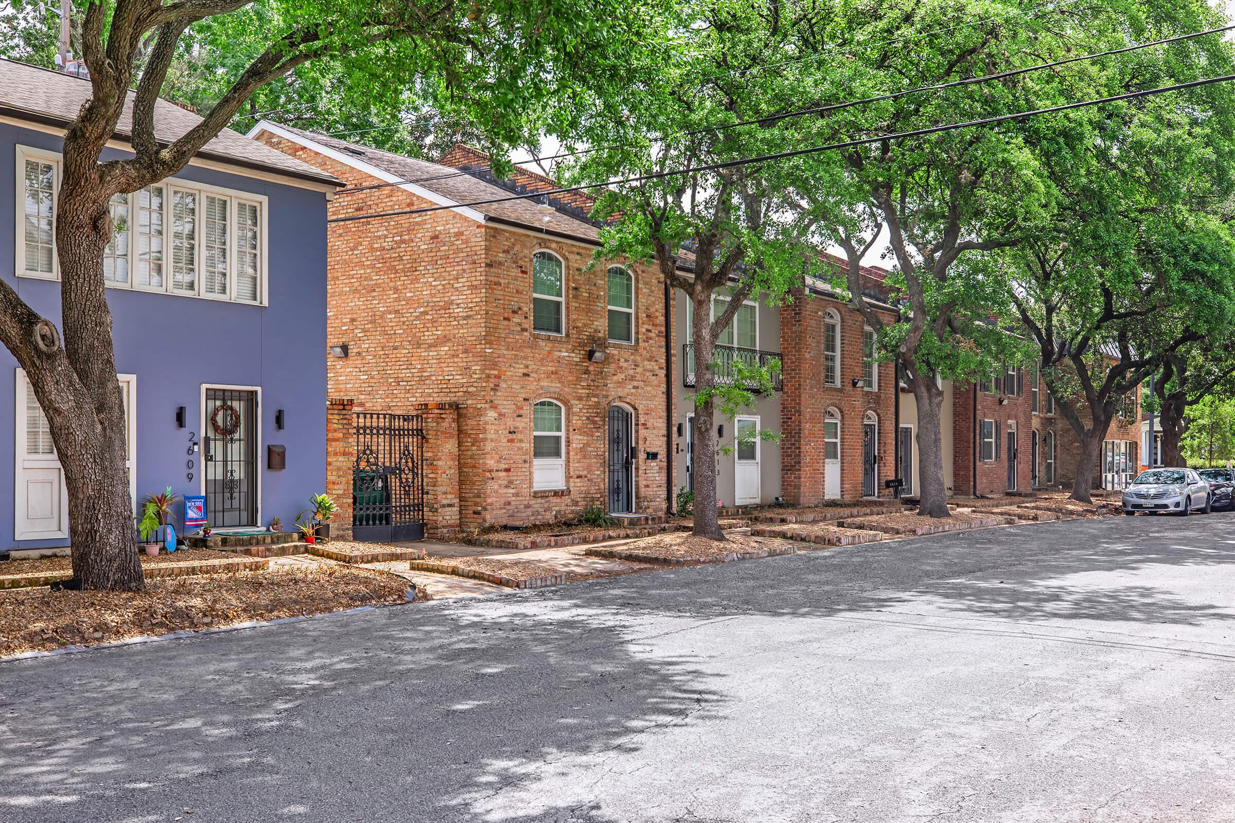 A quiet street scene featuring a row of houses with varied architectural styles, including a blue house and brick facades. Tall trees line the street, providing shade over a gravel pathway. Parked cars are visible along the curb, adding to the residential atmosphere of the neighborhood.