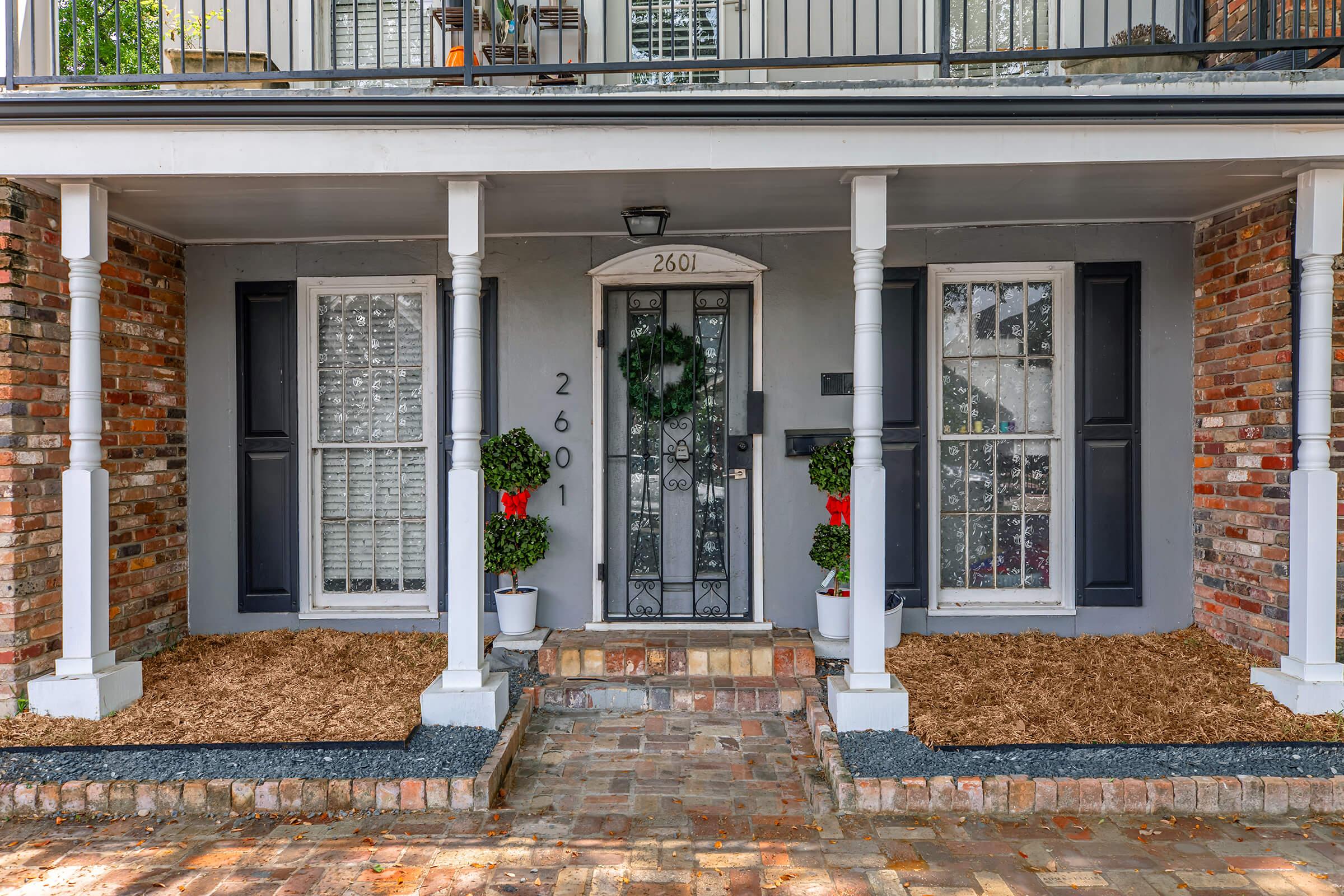 A charming entryway featuring a gray front door with a decorative wreath, flanked by two white windows adorned with festive decorations. The brick exterior is complemented by neatly arranged flower pots and fresh mulch lining the pathway. The house number 2601 is visible above the door.