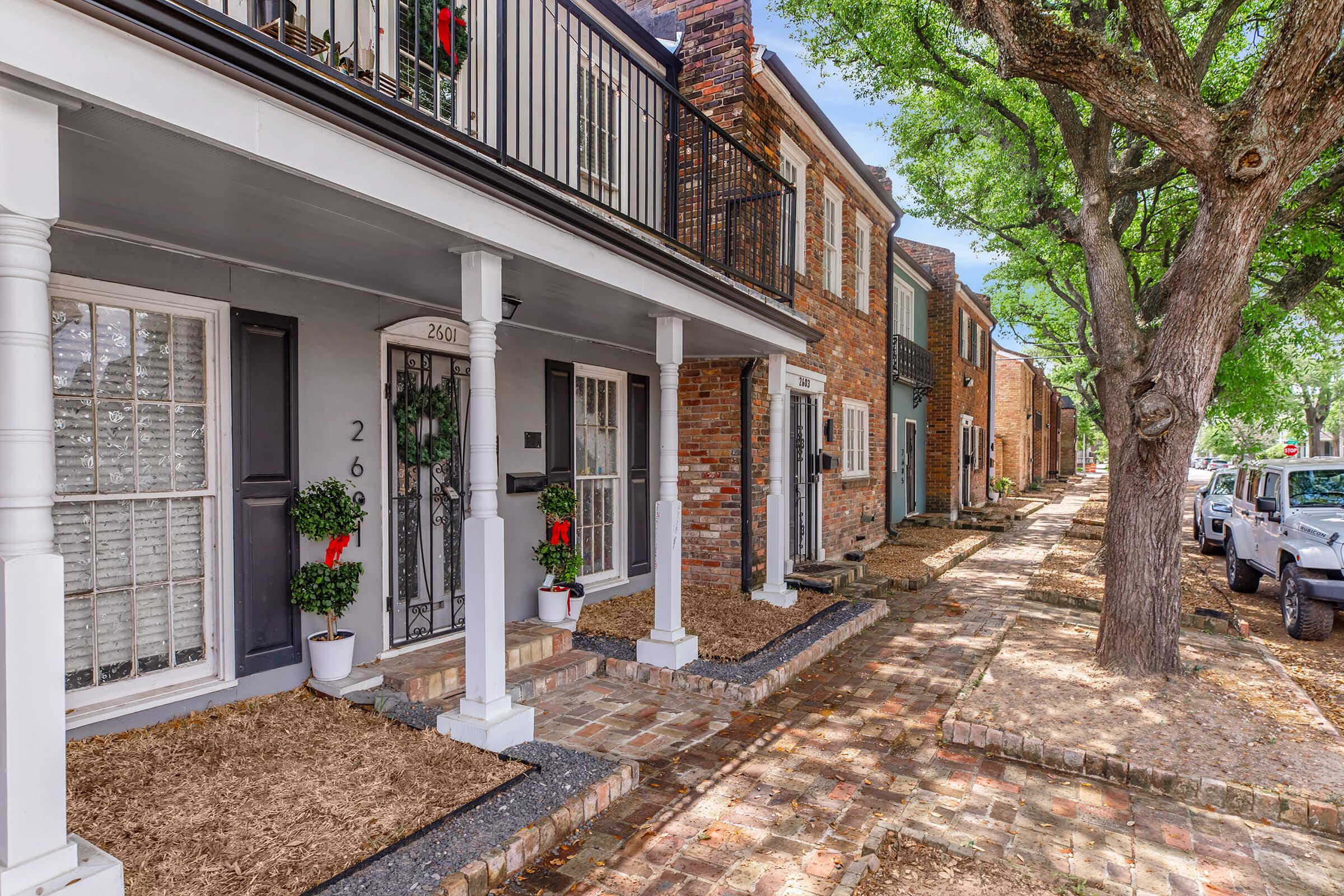 Row of charming residential homes with varied brick exteriors and decorative elements. Each entrance features festive greenery, and the sidewalk is lined with trees. The scene conveys a quaint, inviting neighborhood atmosphere.