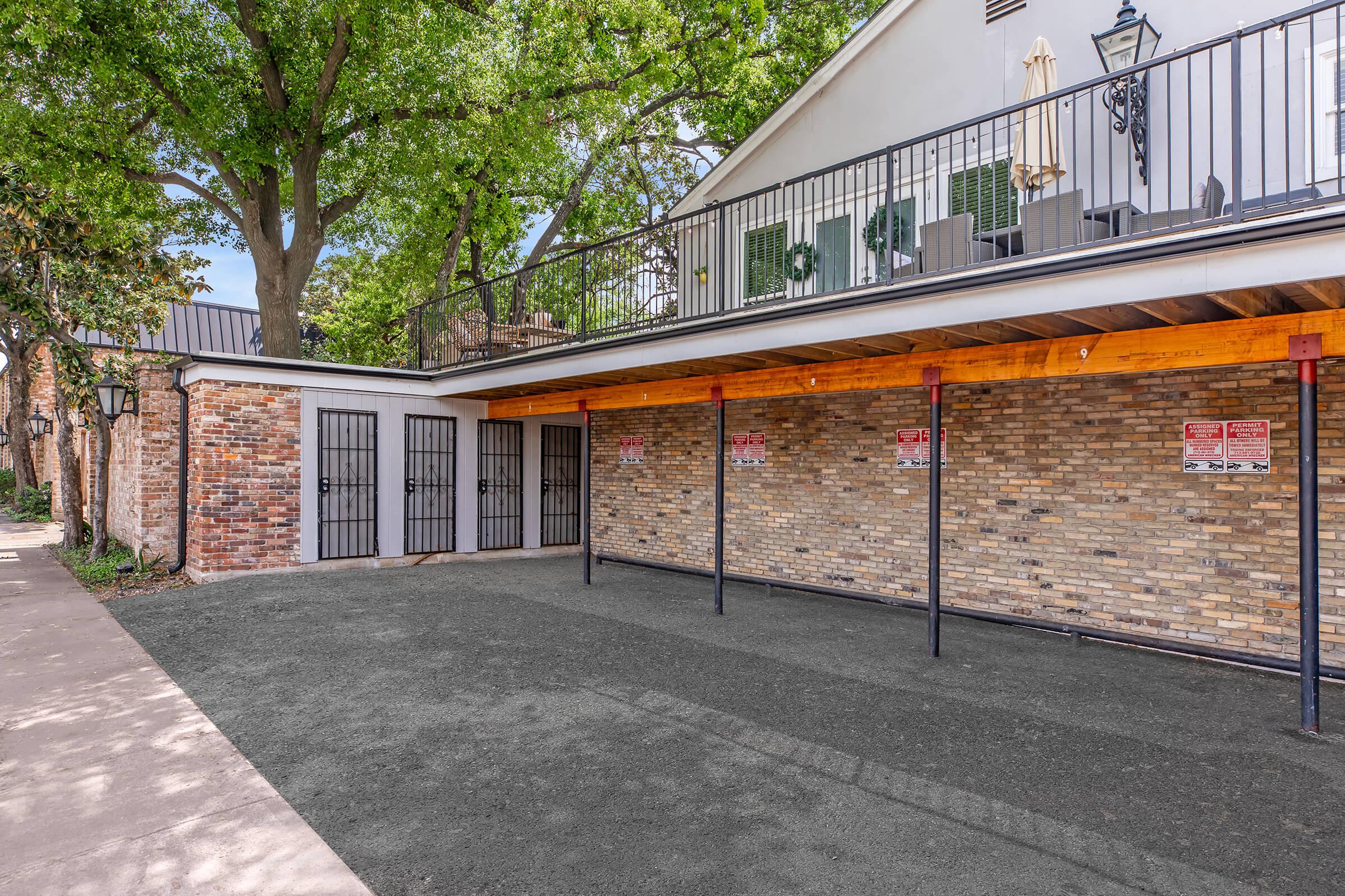 A view of a parking area with a brick wall and wooden beams overhead. There are multiple gated entrances, some with warning signs. The surrounding area features greenery from nearby trees and a glimpse of a building with a second-floor balcony and outdoor seating.