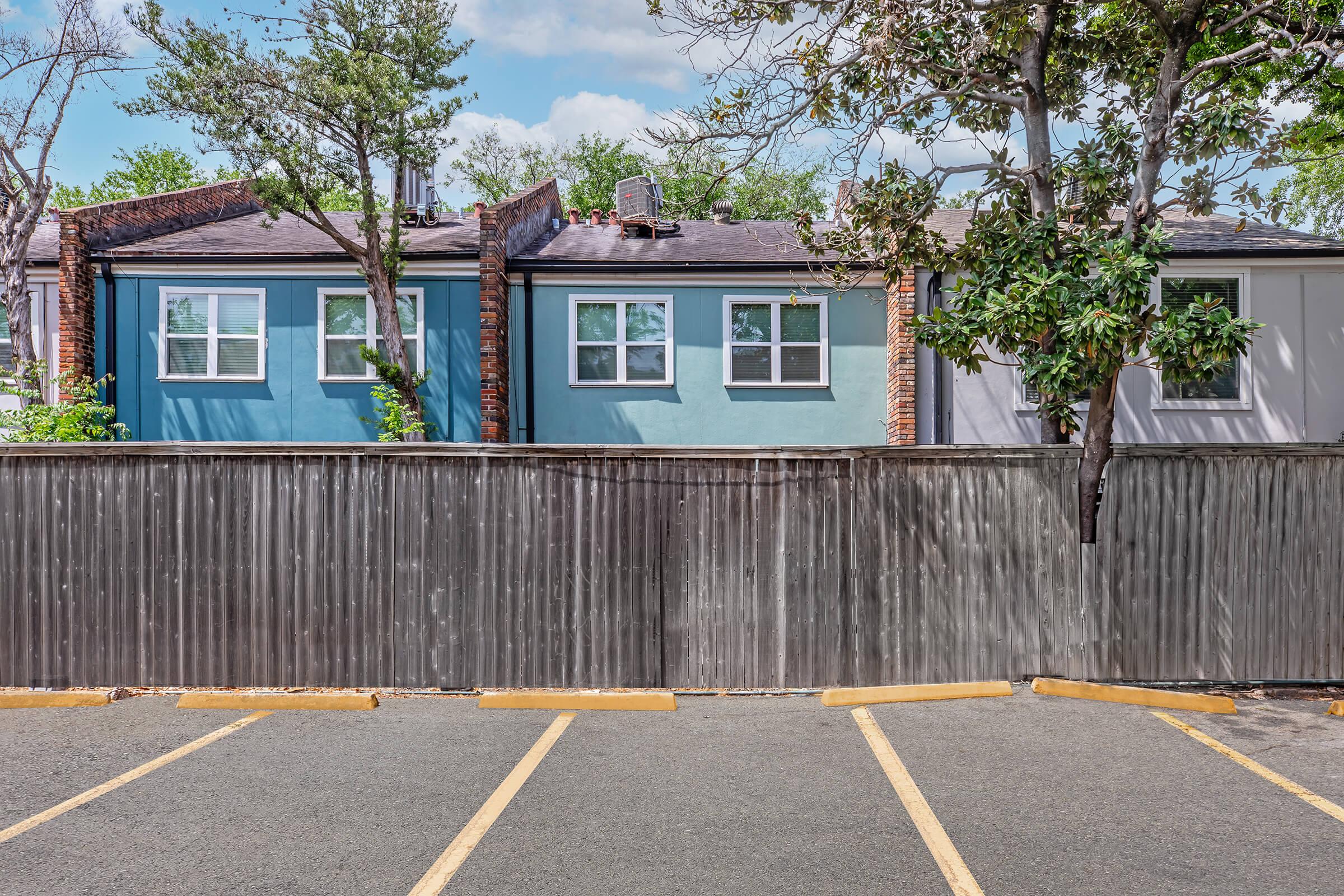 A view of a row of homes with blue and grey exteriors, partially obscured by a wooden fence. The parking lot in front has yellow markings for parking spaces. Lush green trees are visible, and the sky is partly cloudy.