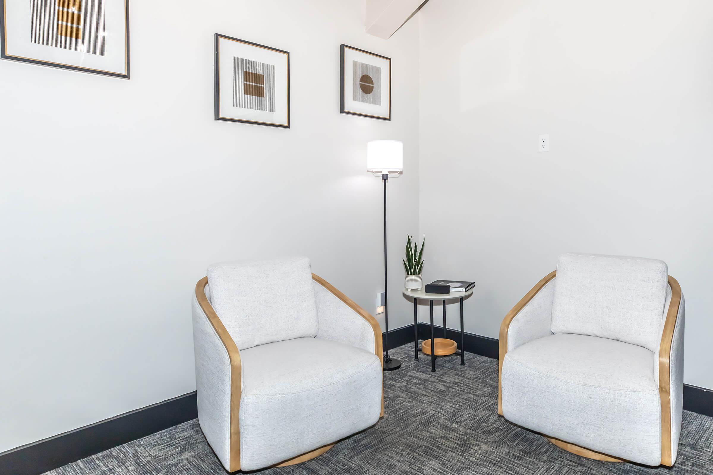 A cozy interior corner featuring two light gray upholstered chairs with wooden frames. A small round black side table stands between them, holding a stack of books and a potted plant. A modern floor lamp provides soft lighting, and three framed artworks adorn the wall above the chairs. The floor is covered with gray carpeting.