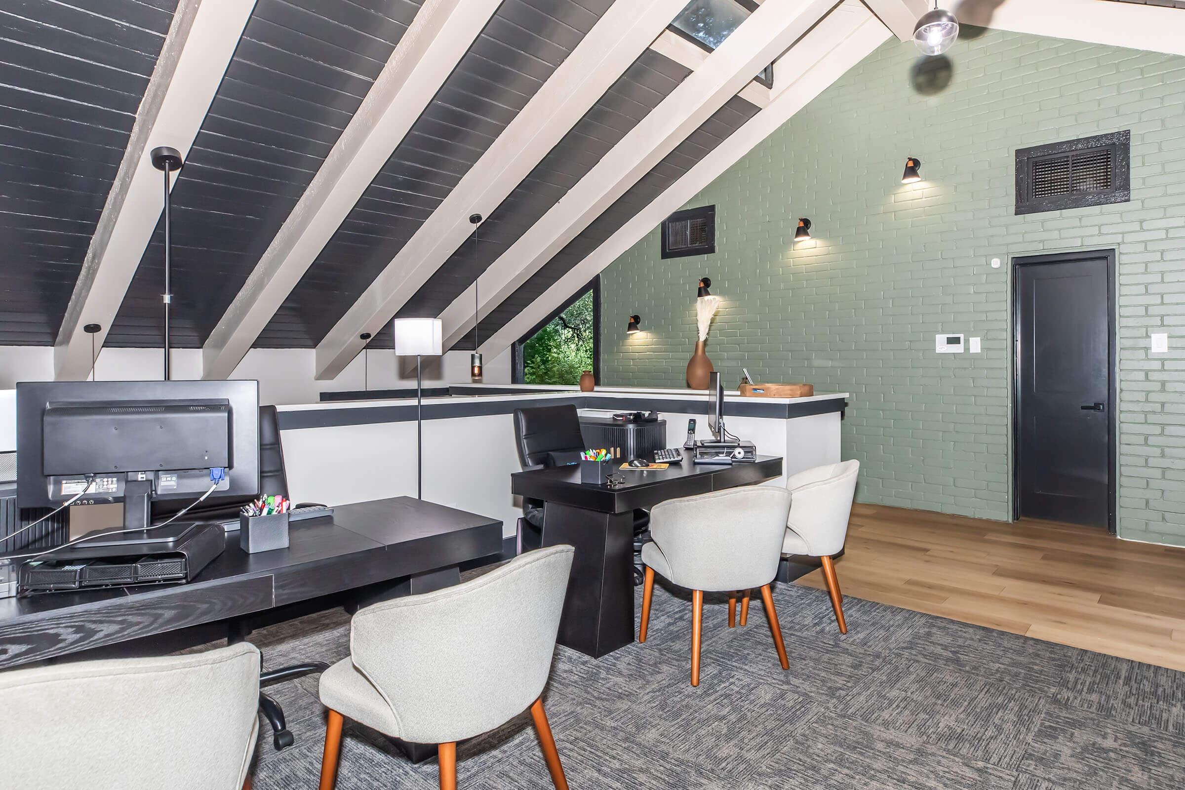 Modern office interior featuring sloped ceilings, large windows, and stylish decor. Two workstations with computer monitors and office supplies are set against a green brick wall. Comfortable chairs are positioned nearby, and there's ambient lighting from wall sconces and a standing lamp. The floor is wood, adding warmth to the space.