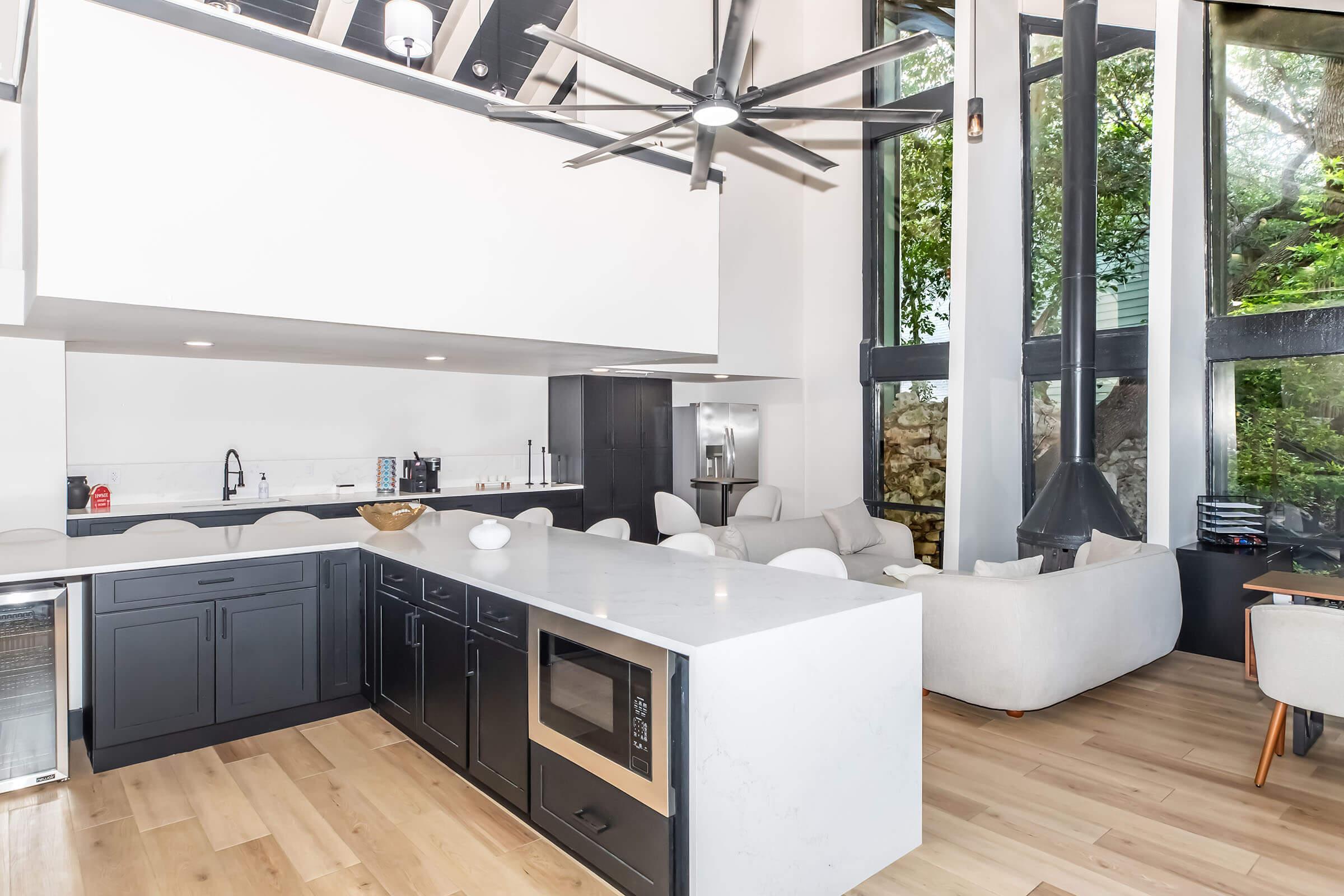 Modern kitchen featuring a large island with a microwave, sleek black cabinetry, and a minimalist design. Large windows allow natural light to flood the space, complemented by a stylish ceiling fan. A cozy living area with soft furnishings is visible in the background, creating an open and inviting atmosphere.