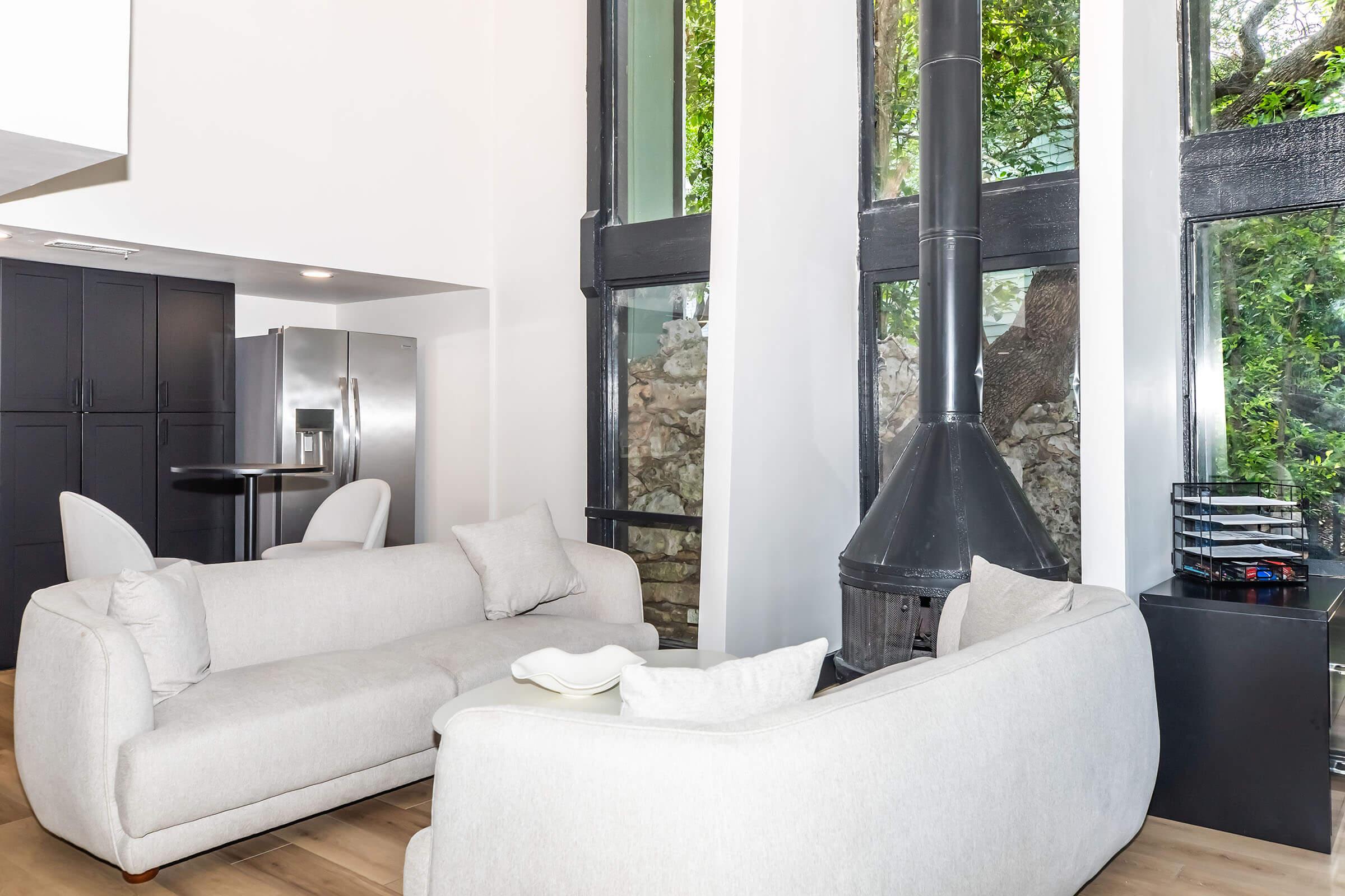 A modern living room featuring two light gray sofas facing each other. There's a sleek black wood stove in the corner, tall windows allowing natural light, and a view of greenery outside. A black shelving unit is visible, along with a refrigerator in the background. The decor is minimalist and inviting.