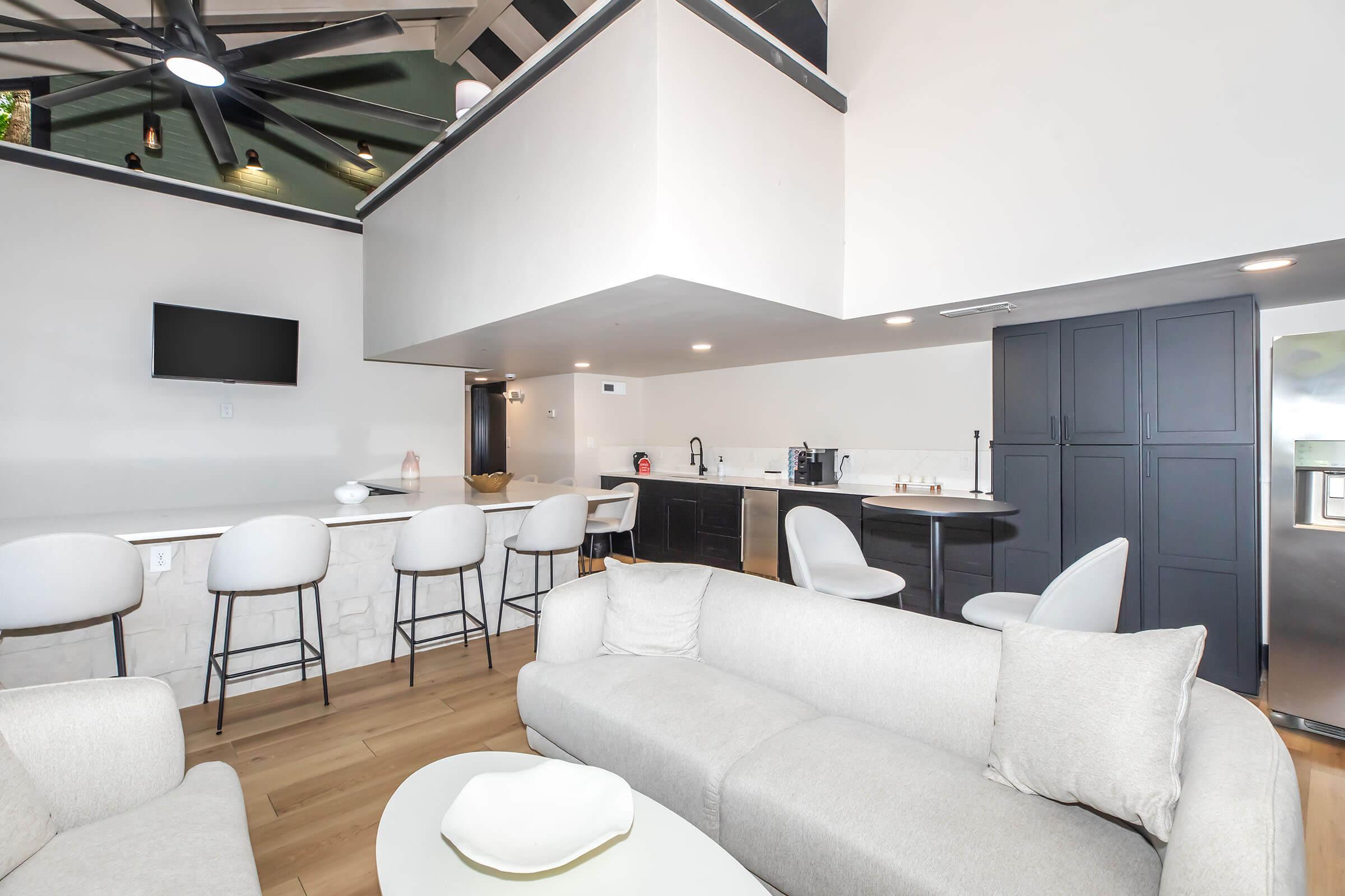 A modern interior of a stylish living space featuring a light-colored sofa, a round coffee table, and a kitchen area with bar stools. The design includes a ceiling fan, a wall-mounted TV, and sleek cabinets, creating an open and inviting atmosphere with warm wooden flooring.