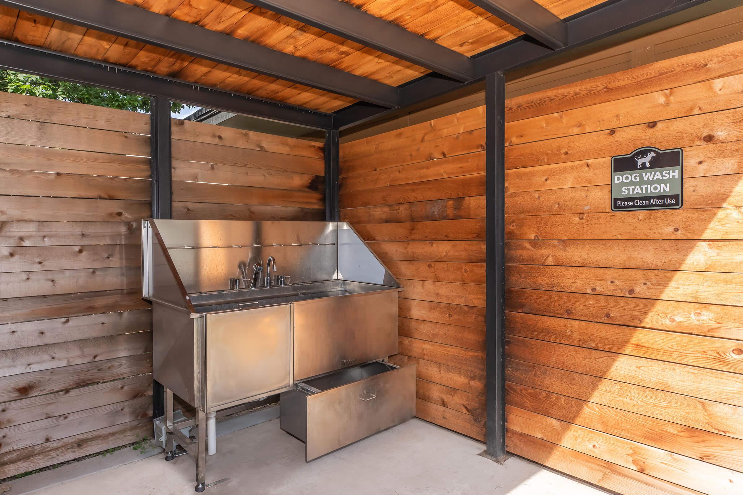 A stainless steel dog wash station enclosed by wooden walls. The station features multiple faucets and a drainage area below. A sign above reads "DOG WASH STATION" with a note to "Please Clean After Use." The space is well-lit, with a roof overhead.