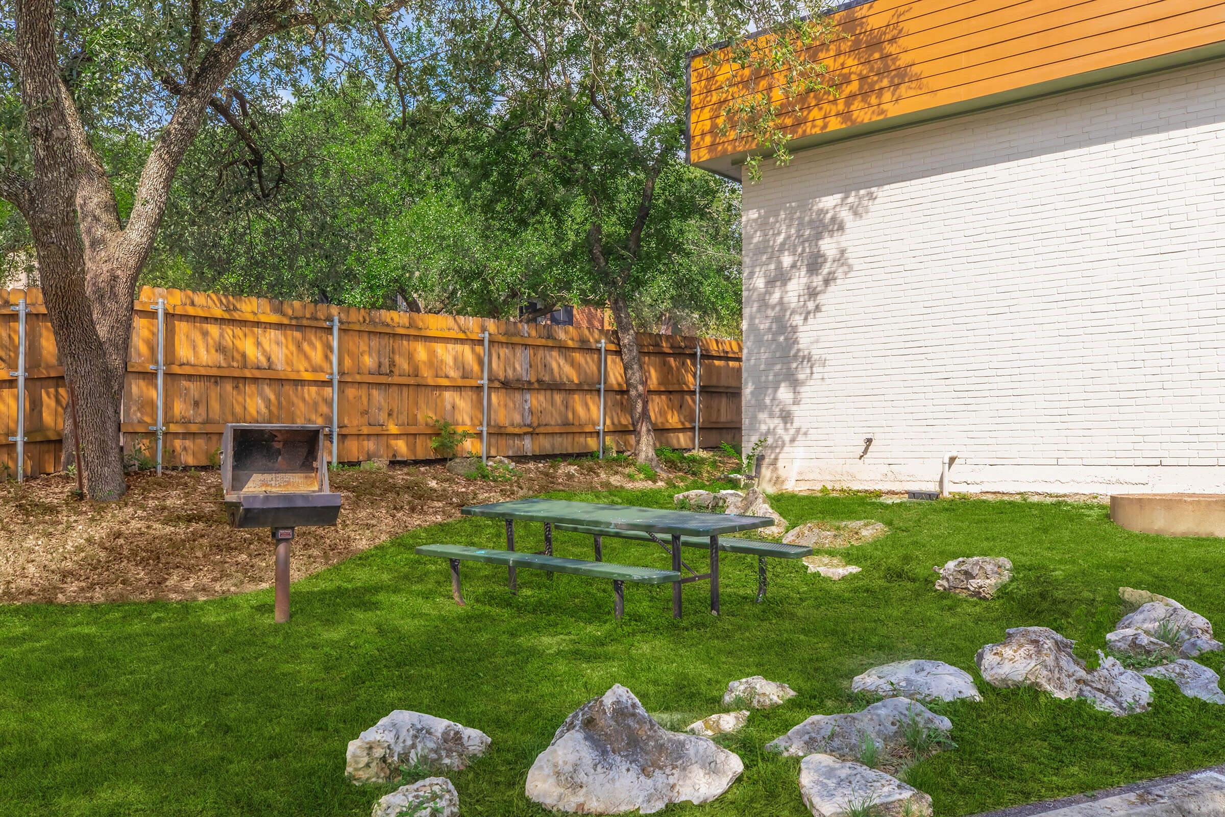 A cozy outdoor space featuring a picnic table on lush green grass, surrounded by large rocks and greenery. There's a barbecue grill nearby, set against a backdrop of a wooden fence and a modern white building with a wooden accent. Sunlight filters through the trees, creating a welcoming atmosphere.
