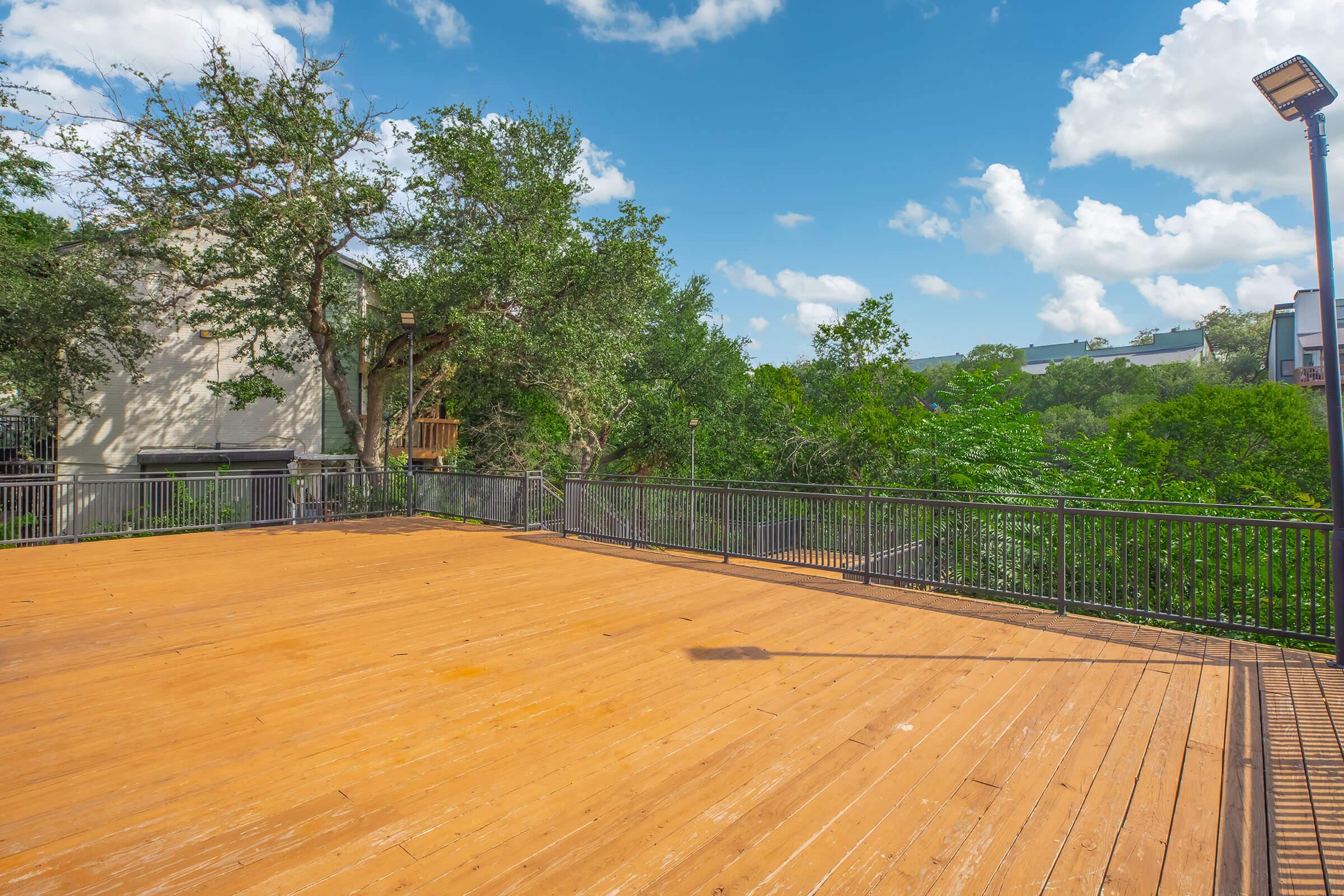 A spacious wooden deck surrounded by lush greenery and trees, with a bright blue sky and fluffy clouds overhead. The scene conveys a tranquil outdoor space suitable for relaxation or gatherings, with a railing providing safety and a hint of modern architecture in the background.