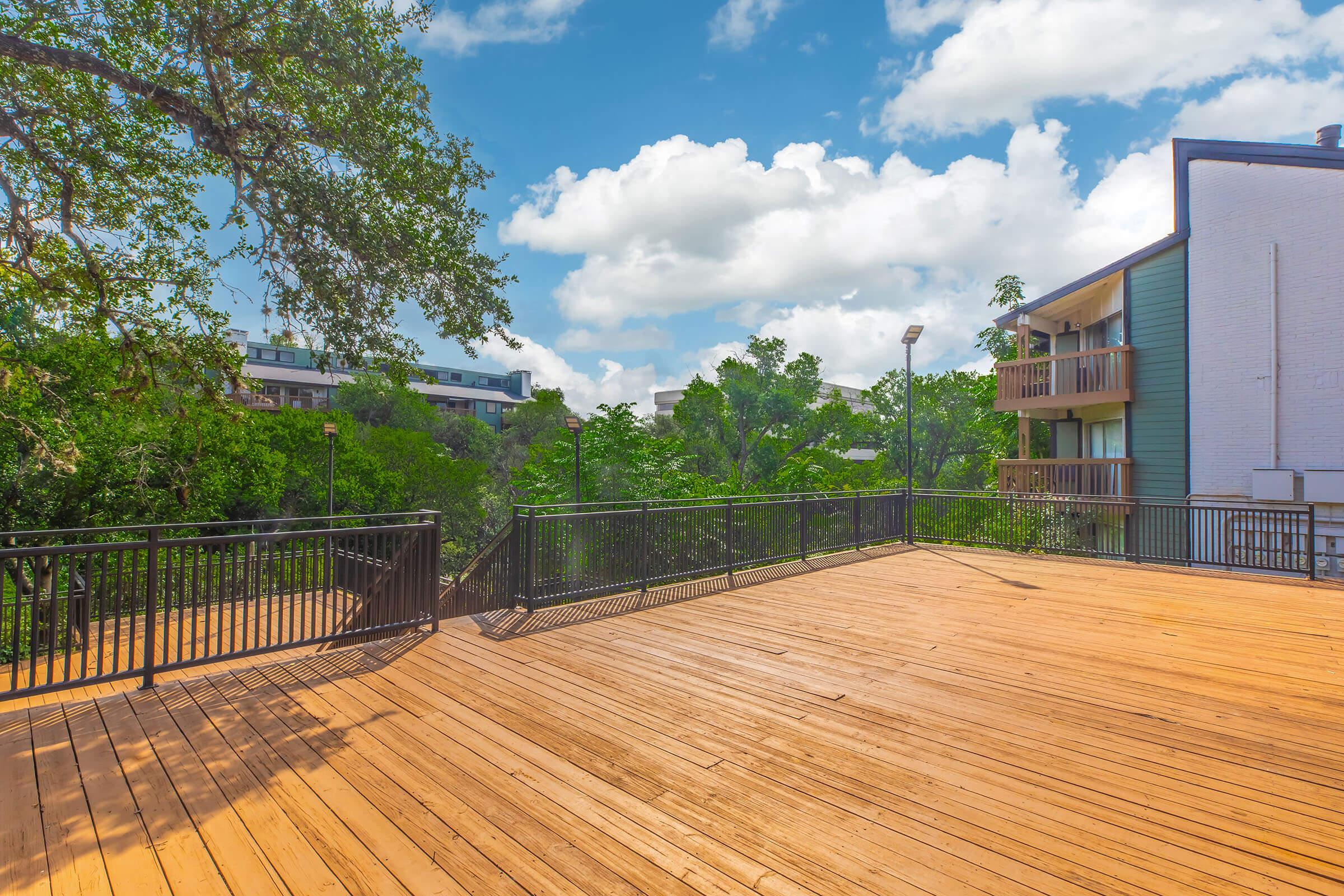 Spacious wooden deck with a black railing, surrounded by lush greenery and trees. In the background, there are residential buildings partially obscured by foliage under a bright blue sky with fluffy white clouds. The scene depicts a tranquil outdoor area suitable for relaxation or gatherings.