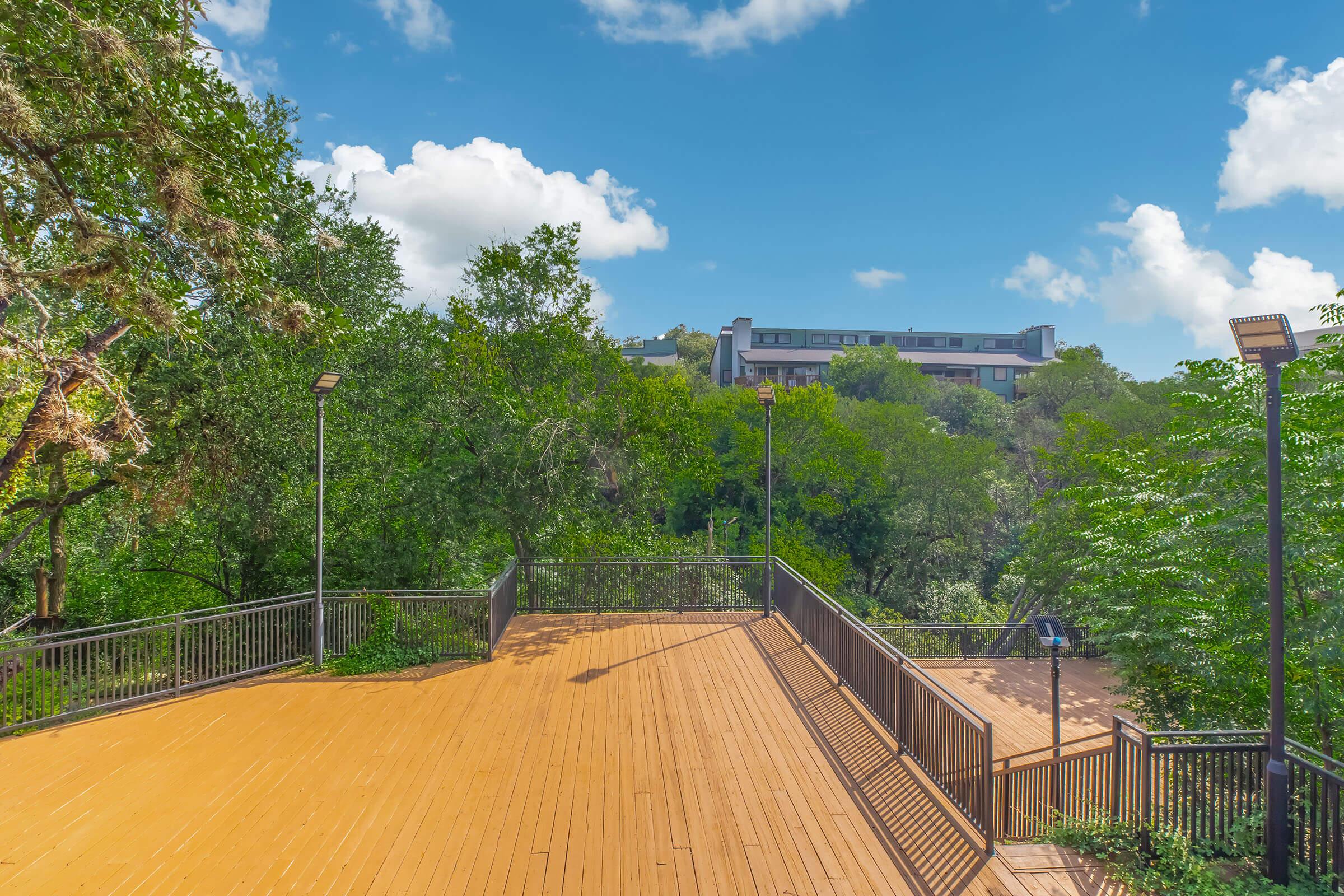 A wooden deck overlooking a lush green landscape with trees and a building in the background under a bright blue sky with fluffy clouds. The scene conveys a peaceful outdoor setting, ideal for relaxation or leisure activities.